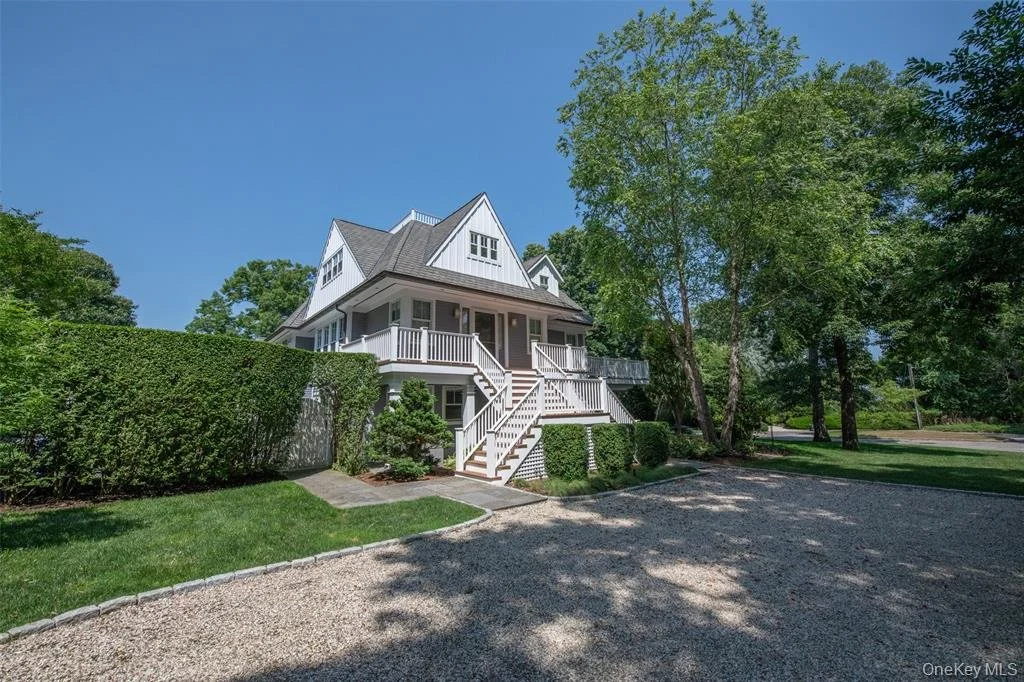 White multi-story house with a steep roof, surrounded by greenery and trees, with a gravel driveway in front.