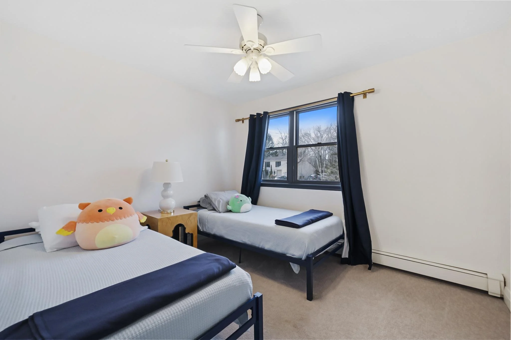A bedroom with three beds, each with plush toys, one resembling a bird and another a green character. The room has white walls, a window with dark curtains, a ceiling fan, and a beige carpet.