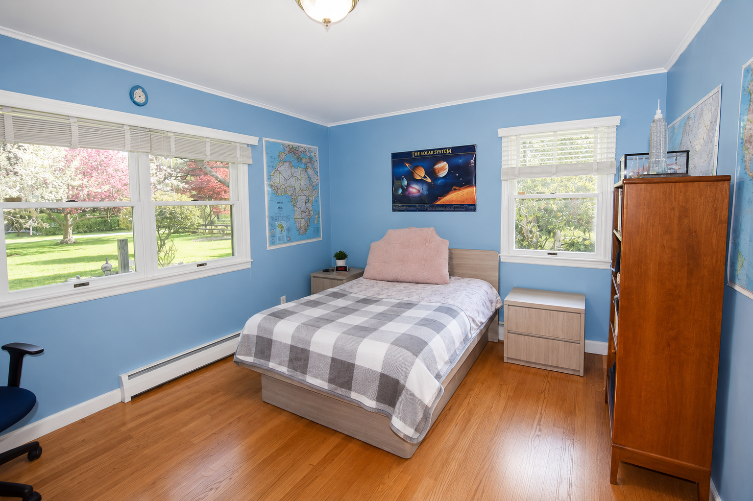 A bedroom with light blue walls, a window showing a garden, a bed with a checkered gray and white blanket, a pink pillow, a nightstand with a small plant and alarm clock, a wooden bookshelf with models and maps, and space-themed posters.