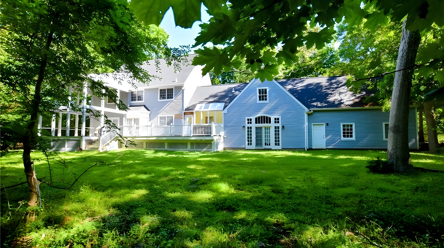 A large, light blue house with a deck and multiple windows, surrounded by green grass and trees.