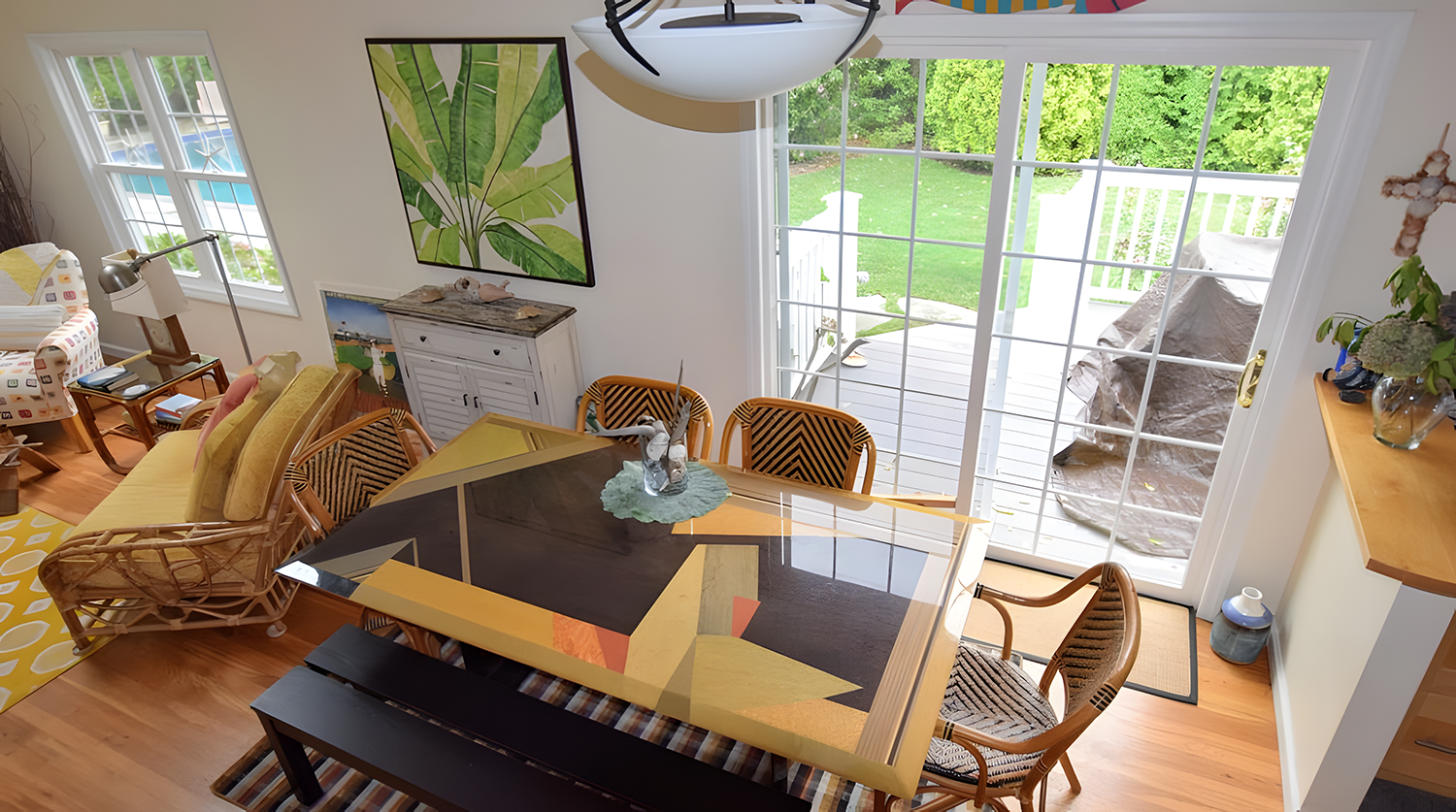 A dining room with a glass sliding door leading to a backyard deck. The room features a rectangular table with a colorful geometric top, surrounded by wooden chairs. There is a rattan bench and an armchair with a yellow cushion, a white sideboard, a 