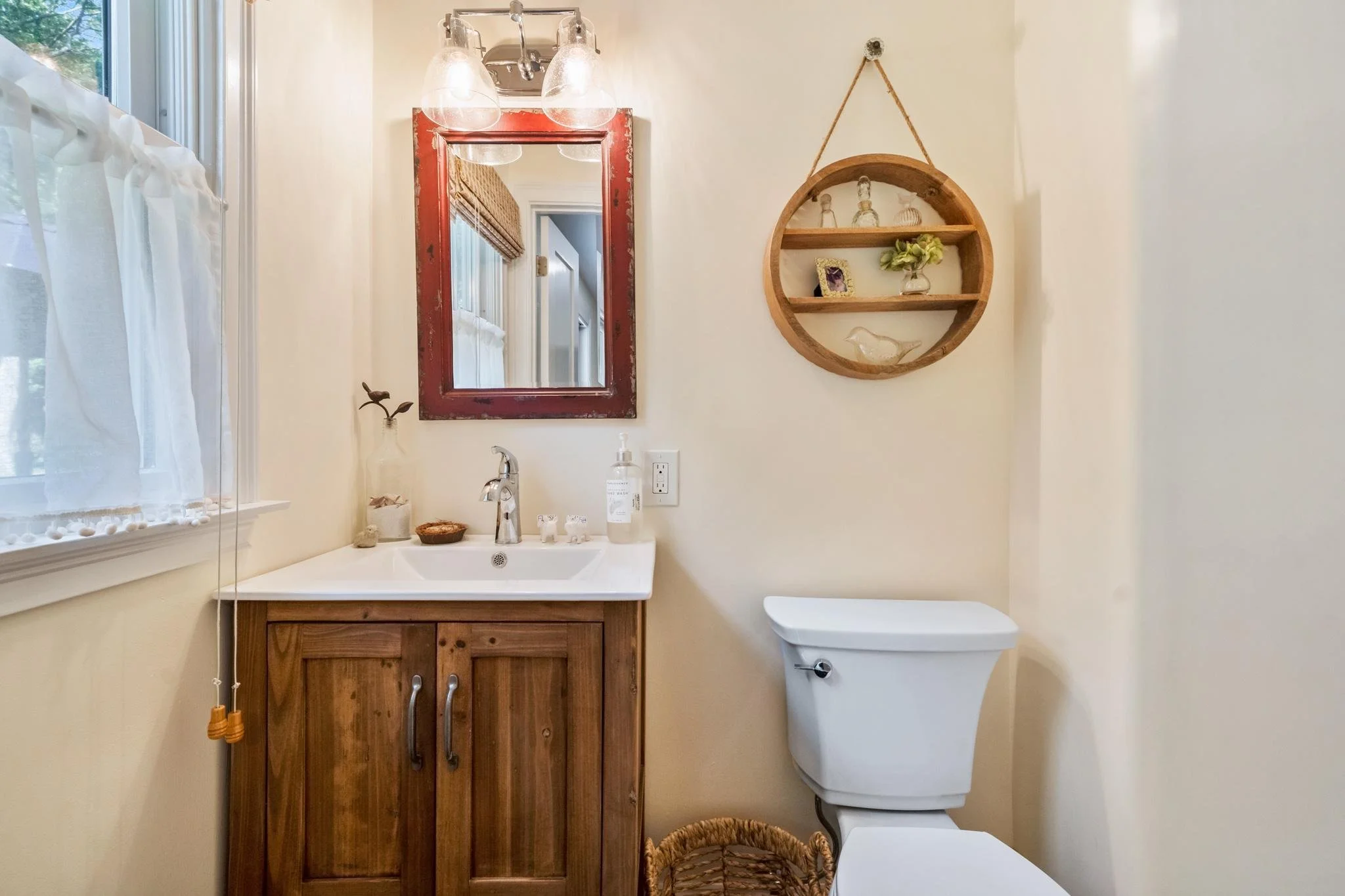 A small bathroom with a window on the left, a rectangular mirror with a red distressed wooden frame above a wooden cabinet with a white sink, a soap dispenser on the counter, and a toilet on the right. A round wooden wall shelf with decorative items 