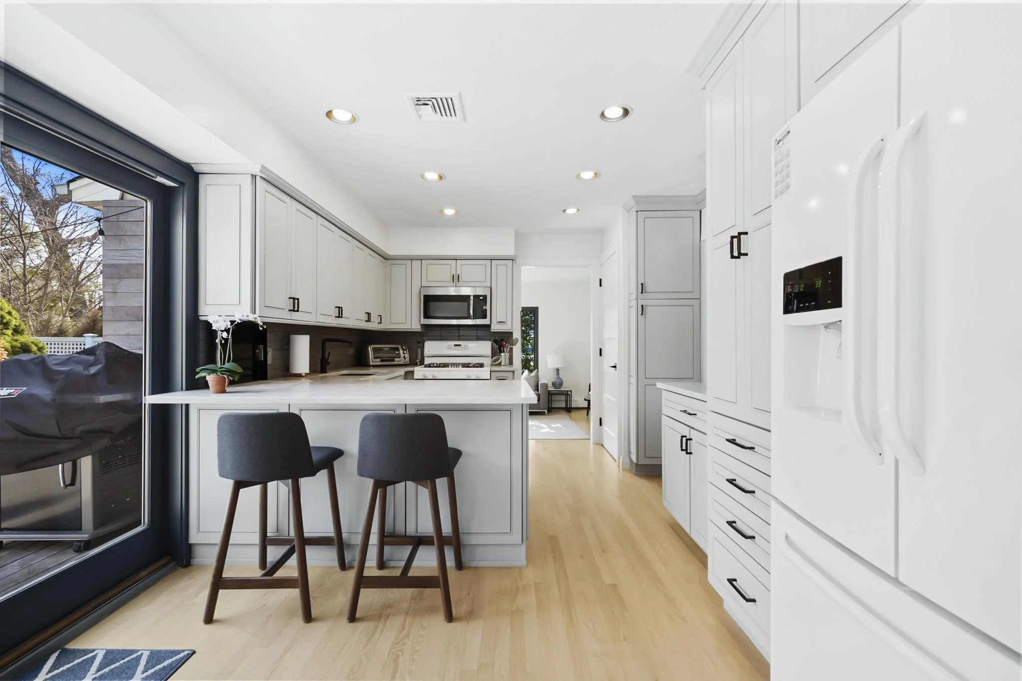Modern white kitchen with built-in appliances, light wood flooring, and a large sliding glass door leading to an outdoor patio. There are two sky chairs at a kitchen island.