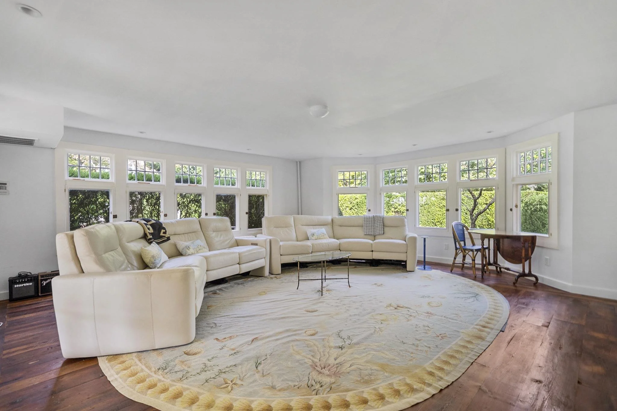Spacious living room with cream-colored sectional sofa, large windows with greenery outside, and a wooden dining table with chairs in the corner.