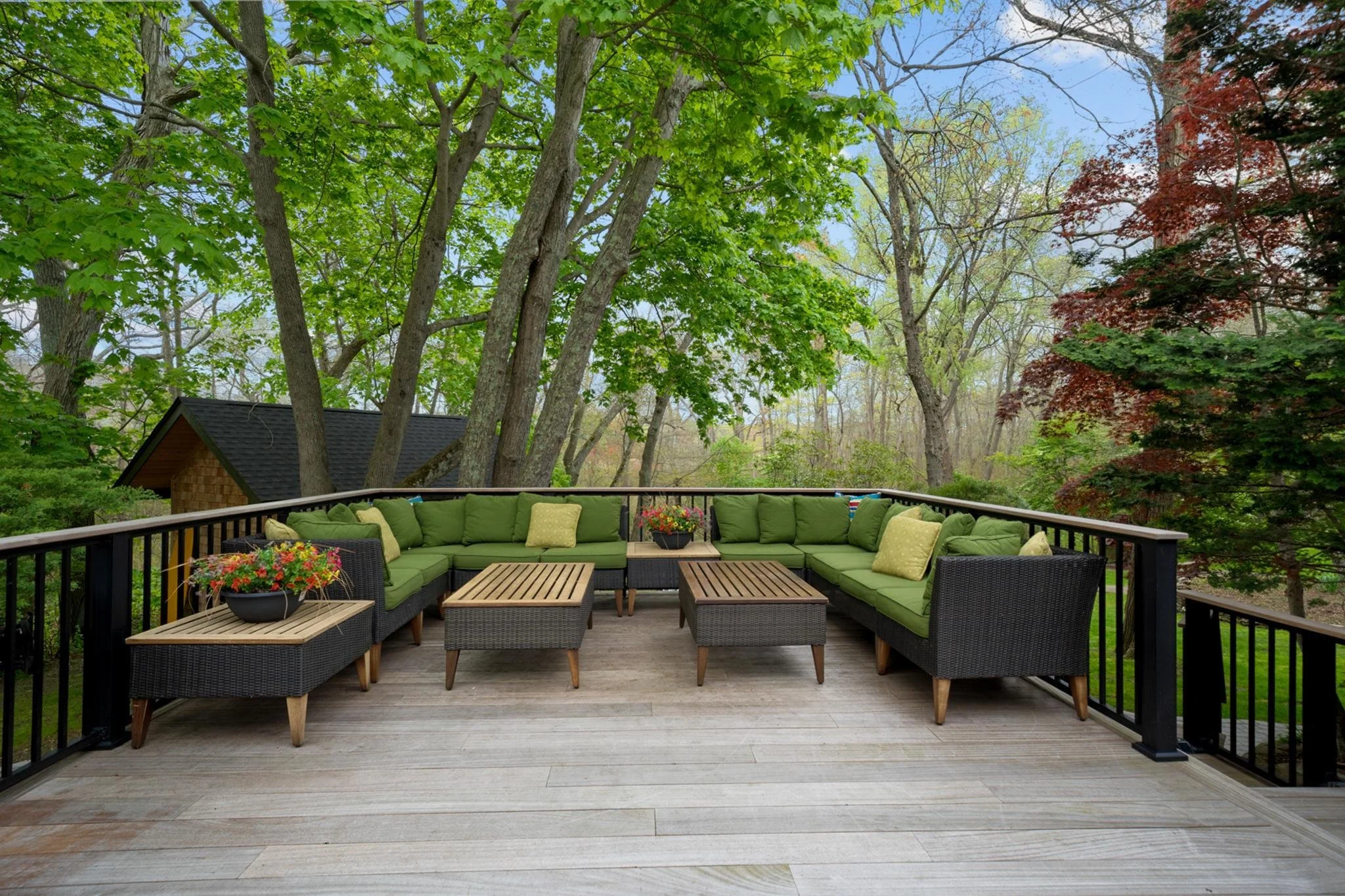 An outdoor deck with green cushioned seating arranged in a U-shape, coffee tables, and potted plants, surrounded by large trees with green and red leaves.