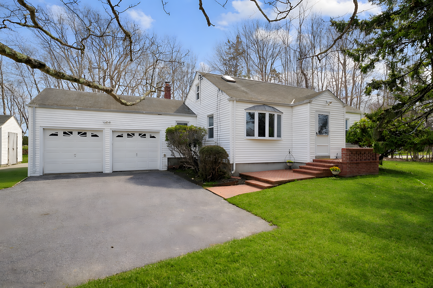 A white house with attached garage, brick steps leading to front door, green lawn, trees in background, blue sky with some clouds.