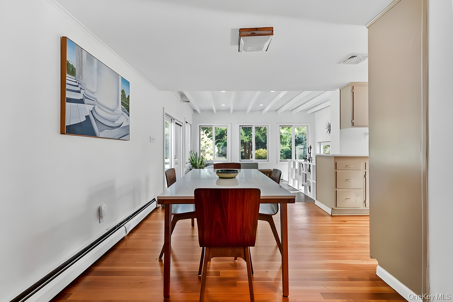 Dining room with wooden table and chairs, large windows with trees outside, artwork on white wall, wooden floors, and ceiling beams.