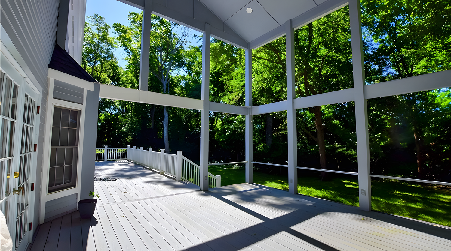View of a spacious outdoor screened porch with white flooring, white railings, and support beams, overlooking a green backyard filled with trees and grass, with sunlight casting shadows.