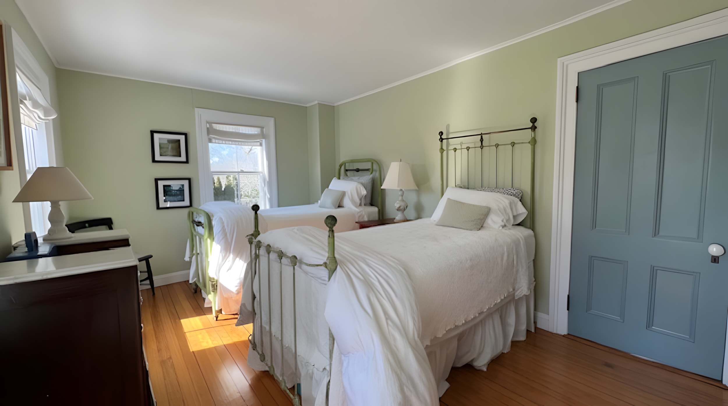 A cozy bedroom with two twin metal beds, white bedding, and pillows. The room has green walls, hardwood floors, and a blue door. There are windows with white blinds, framed artwork on the wall, and a dresser with a lamp and phone.