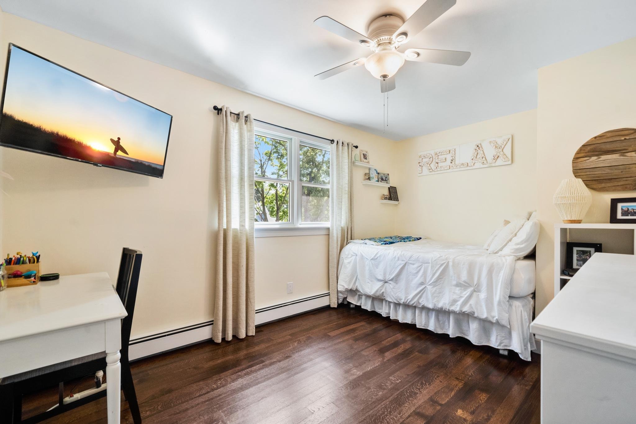 A bedroom with white walls, a ceiling fan, a window with beige curtains, a bed with white bedding, a wall sign that says 'RELAX', a lamp, a picture frame, and other small decor items.