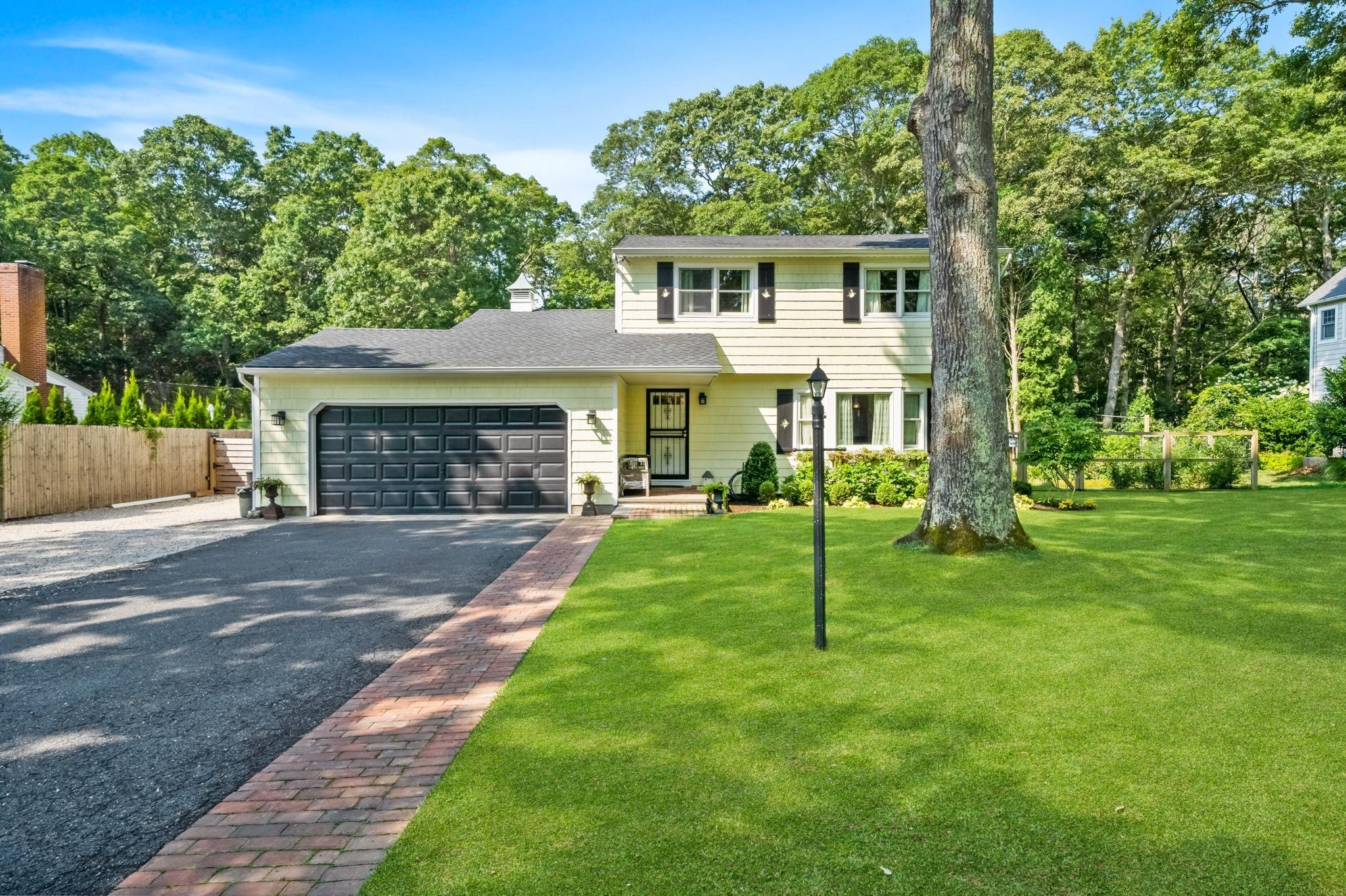 Front view of a two-story yellow house with black shutters, a black garage door, and a brick walkway leading to the front door. The yard has green grass, a large tree, a black lamp post, and a wooden fence on the left. Trees and other houses are in the background on a clear sunny day.