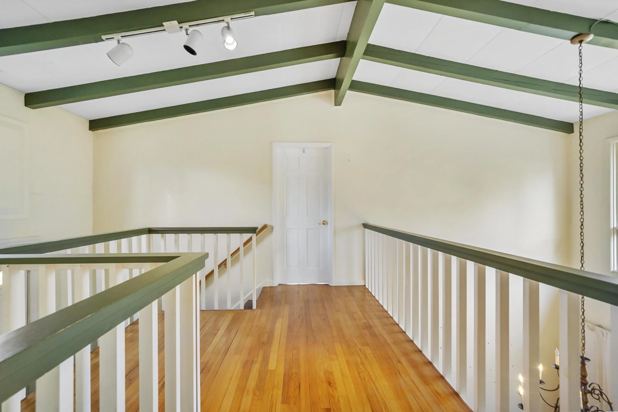 Interior image of a room with a high ceiling supported by green beams and a white ceiling with lighting fixtures. The room has white railing on the sides and a closed white door at the far end. The floor is wooden.