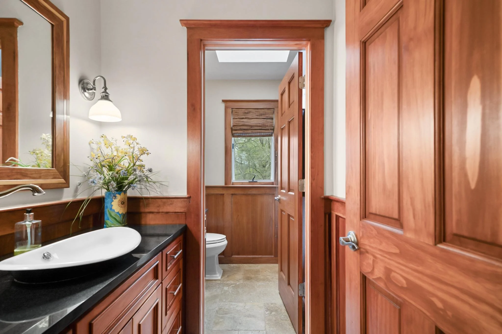 A small bathroom with wooden cabinets, a black countertop, a white sink with a silver faucet, a flower vase, a mirror, a wall-mounted light, a window with blinds, and a toilet in the background.