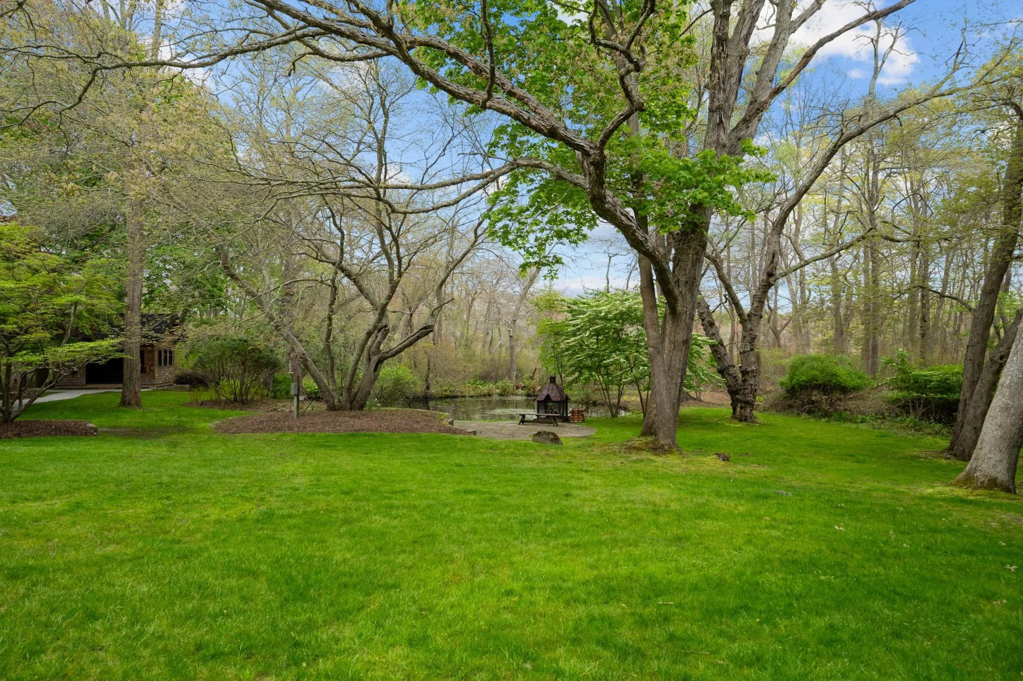 A lush green backyard with well-maintained grass, tall trees with some leaves and some bare branches, a small pond, and a picnic table with a gazebo. There is a house partially visible in the background, with a cloudy sky overhead.