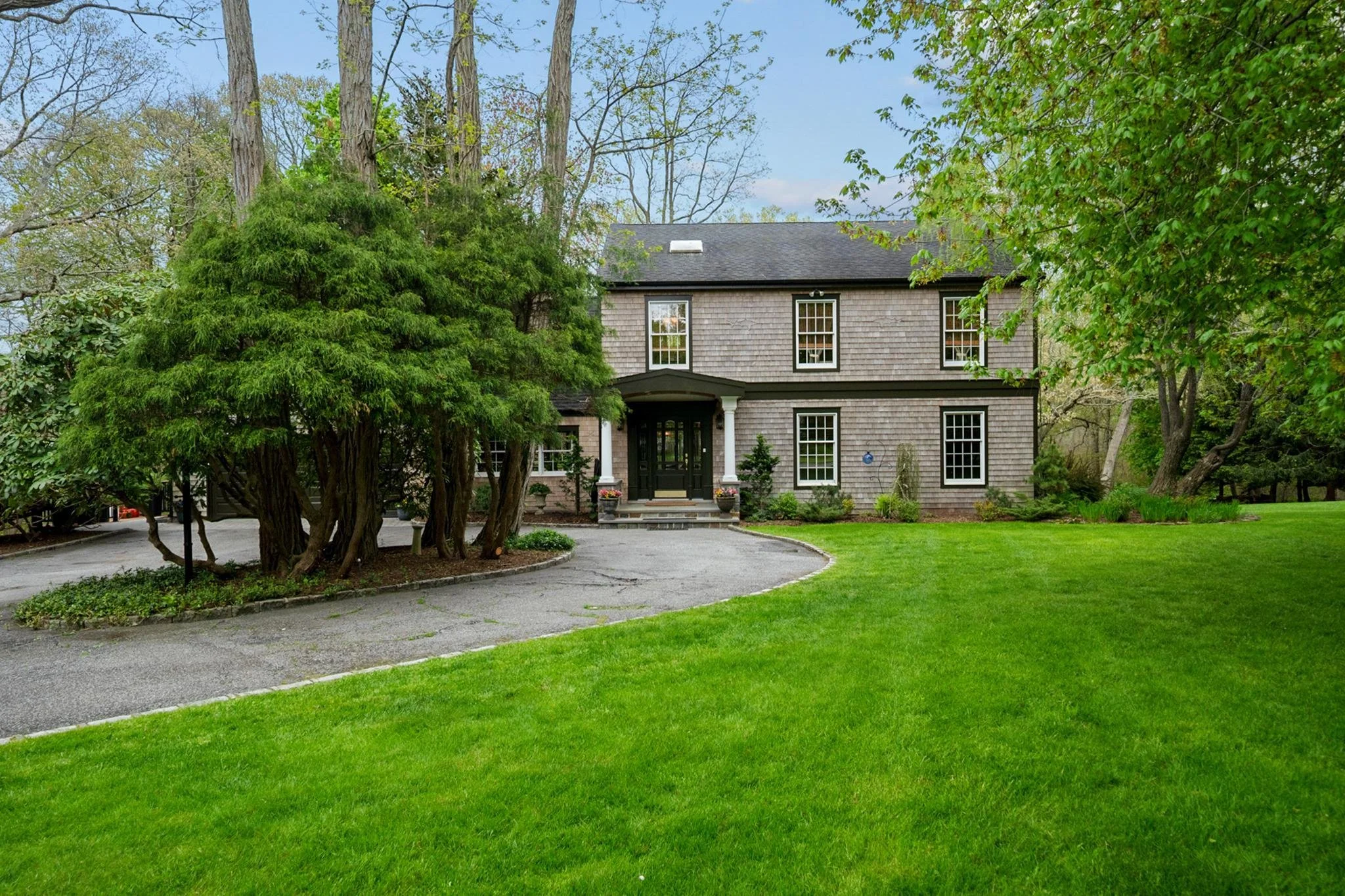 A two-story house with shingle siding, black front door, and white trim, surrounded by green trees and a well-maintained lawn.