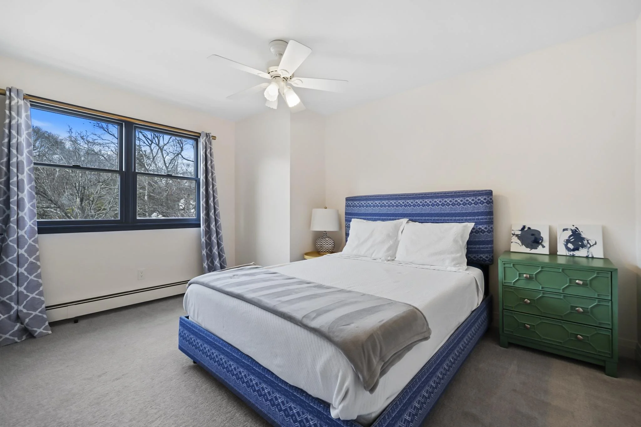 Bedroom with beige walls, a bed with a blue patterned headboard, white bedding, and a gray and white striped blanket. There are two windows with gray and white patterned curtains, a small yellow bedside table with a white lamp, a green dresser, and a
