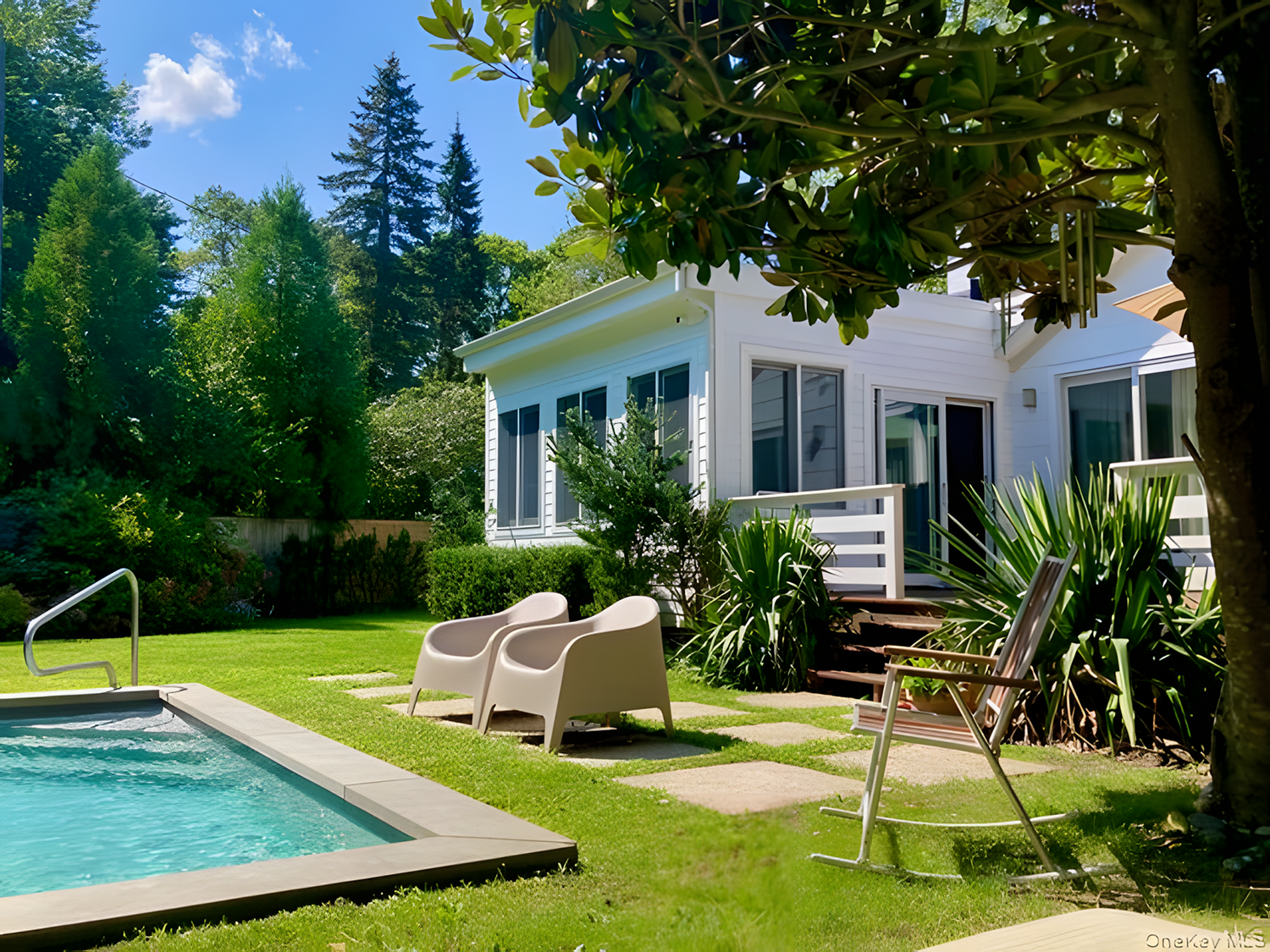 A backyard with a swimming pool, lounge chairs, and a white house with large windows surrounded by green trees and bushes, under a partly cloudy sky.
