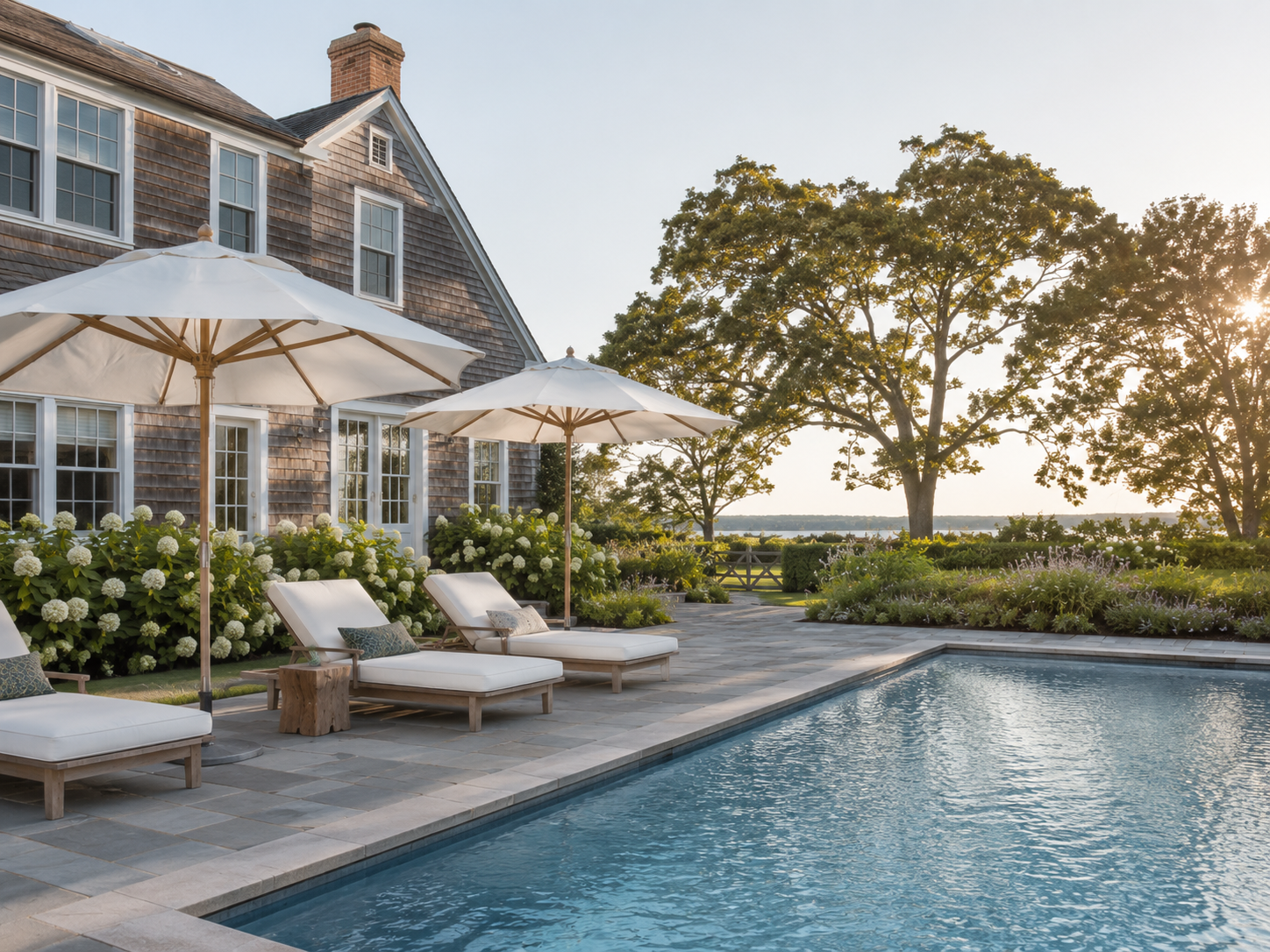 A backyard with a swimming pool, lounge chairs with cushions and umbrellas, surrounded by greenery and flowering bushes, near a large house with a shingled exterior and large windows, with trees and a view of water in the background, during sunset.