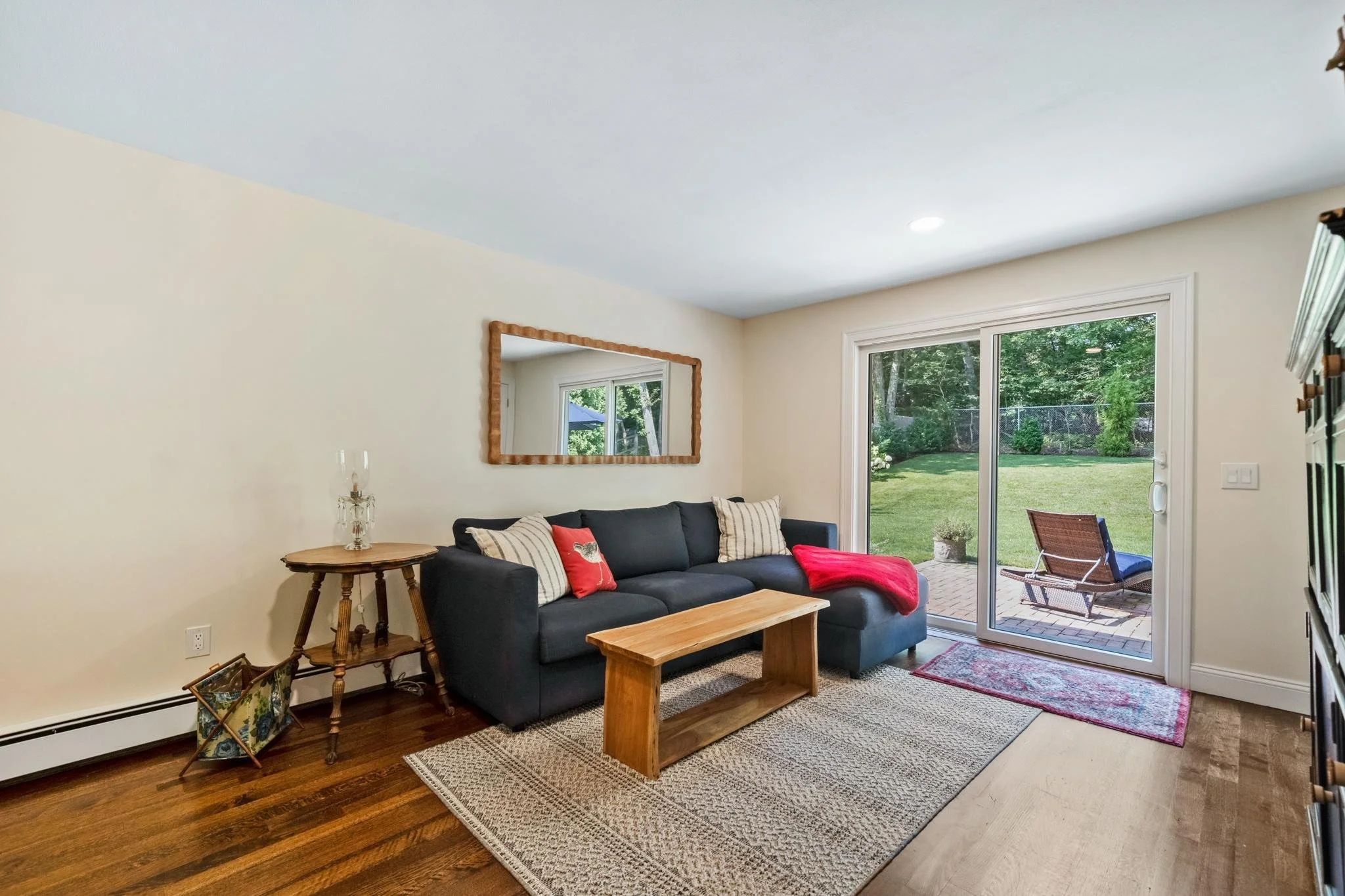 Living room with a blue sofa, striped and red pillows, a wooden coffee table, and a large sliding glass door leading to a backyard patio with outdoor chairs and lush greenery.