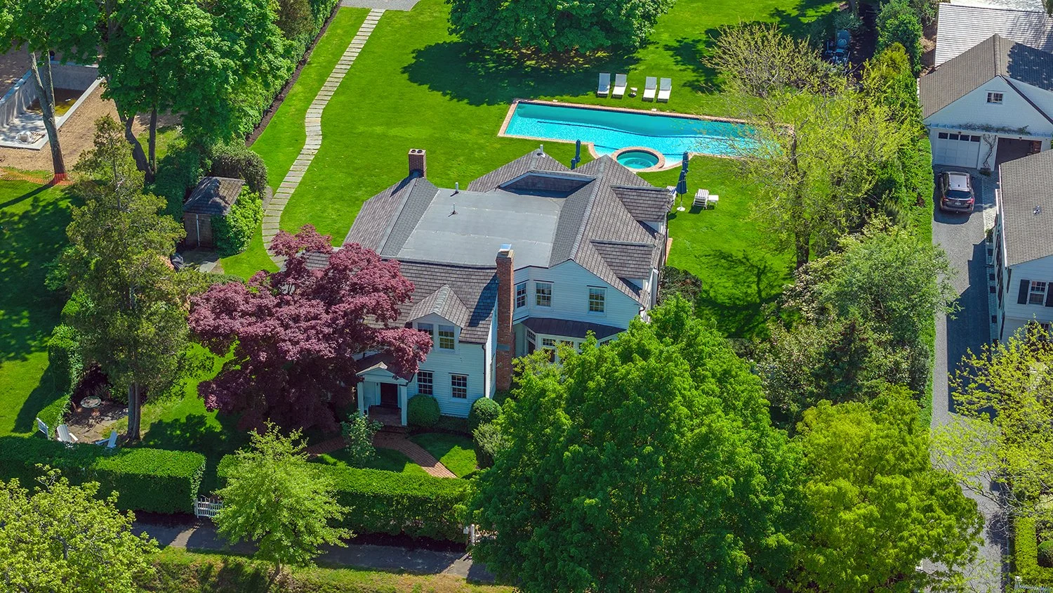 Aerial view of a residential backyard with a swimming pool, hot tub, lush green lawn, mature trees, and a house with a gray roof.