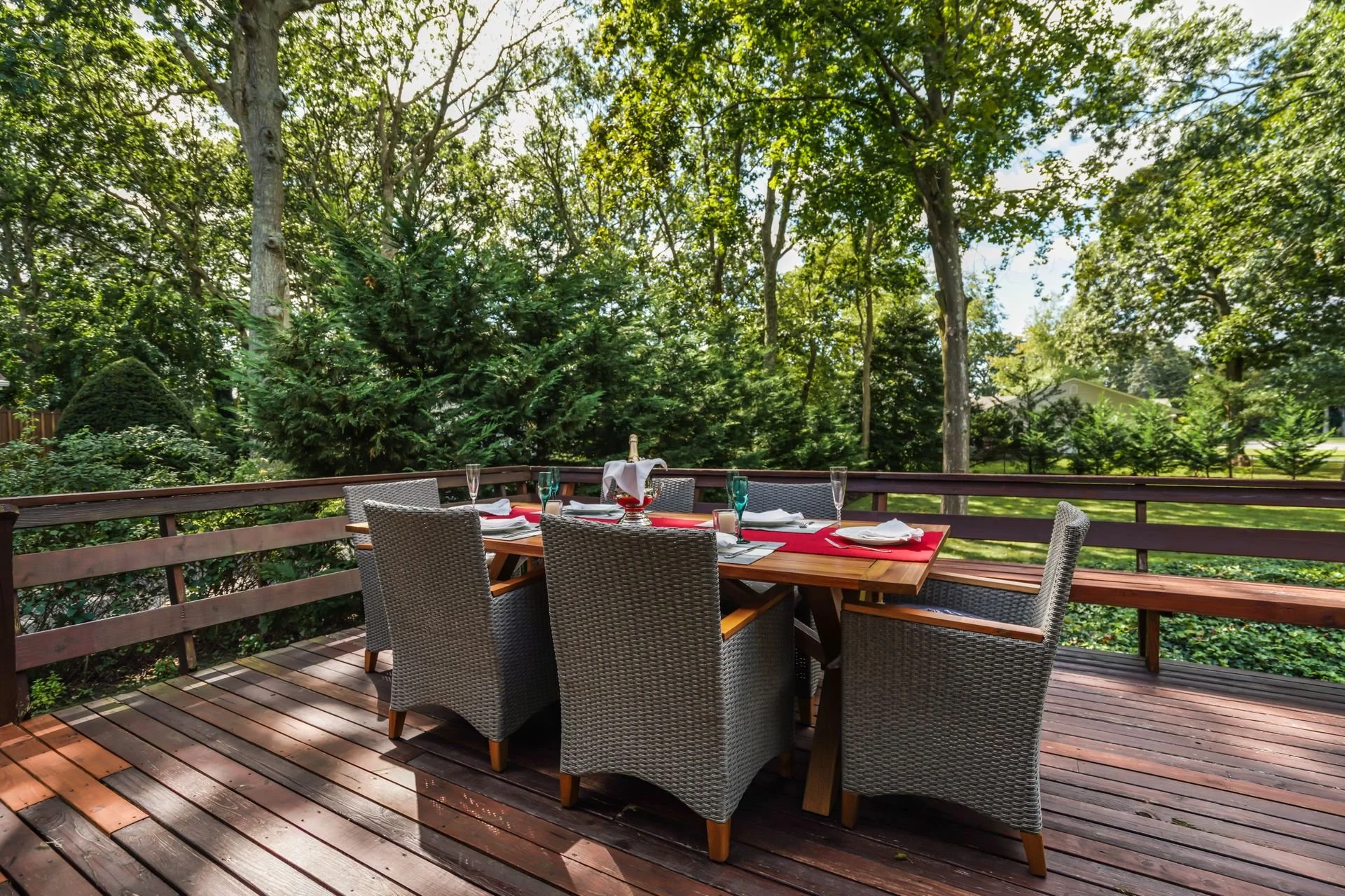 Outdoor dining setup on a wooden deck surrounded by lush green trees, with a dining table set with white plates, glasses, napkins, and a bottle of champagne in an ice bucket.