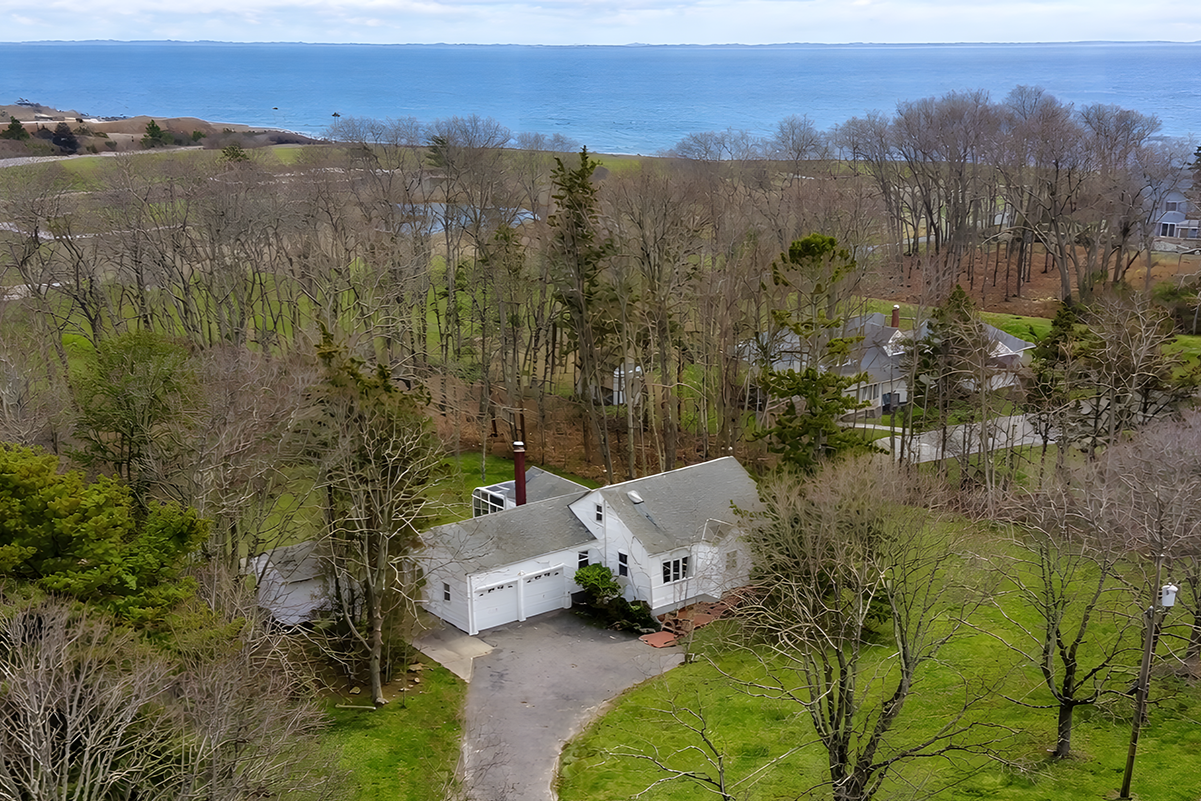 Aerial view of a white house with a gray roof, surrounded by trees and green lawn, overlooking a large body of water in the background.