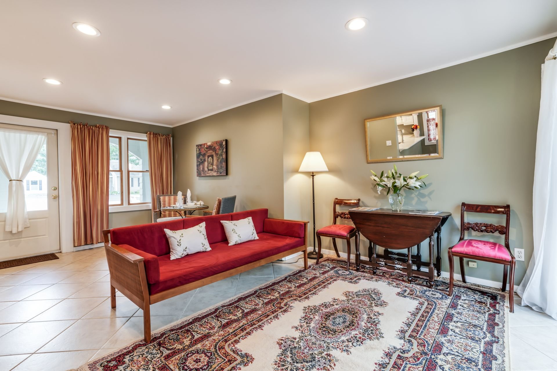 Living room with a red sofa, wooden chairs, a table with a vase of white lilies, framed wall art, a mirror, and a lamp, with windows and curtains, and a patterned rug.