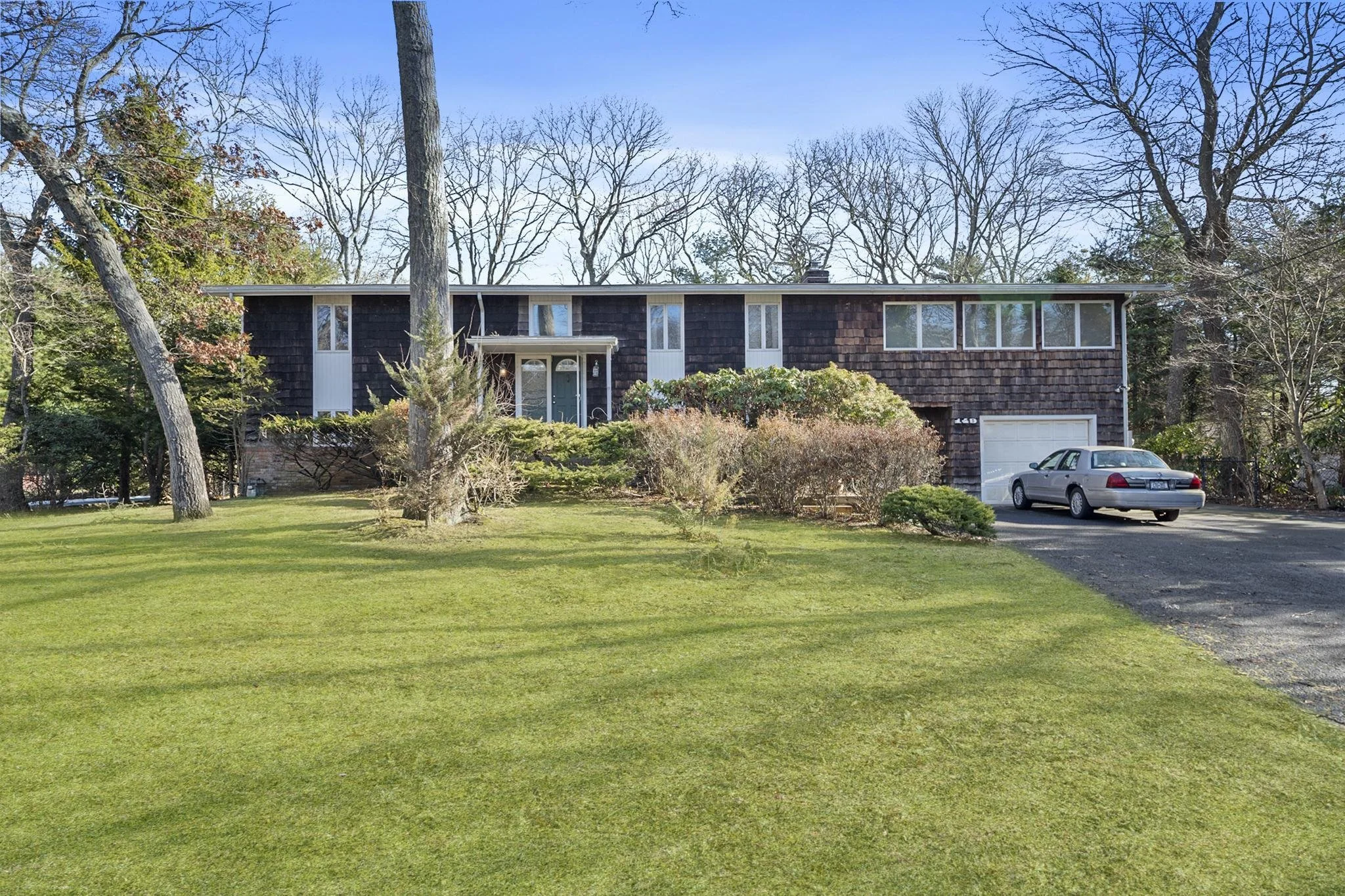 A suburban house with a dark wooden exterior, multiple windows, a white garage door, and a car parked in the driveway. The front yard has a well-maintained green lawn with trees and bushes, and the sky is clear with some leafless trees in the backgro