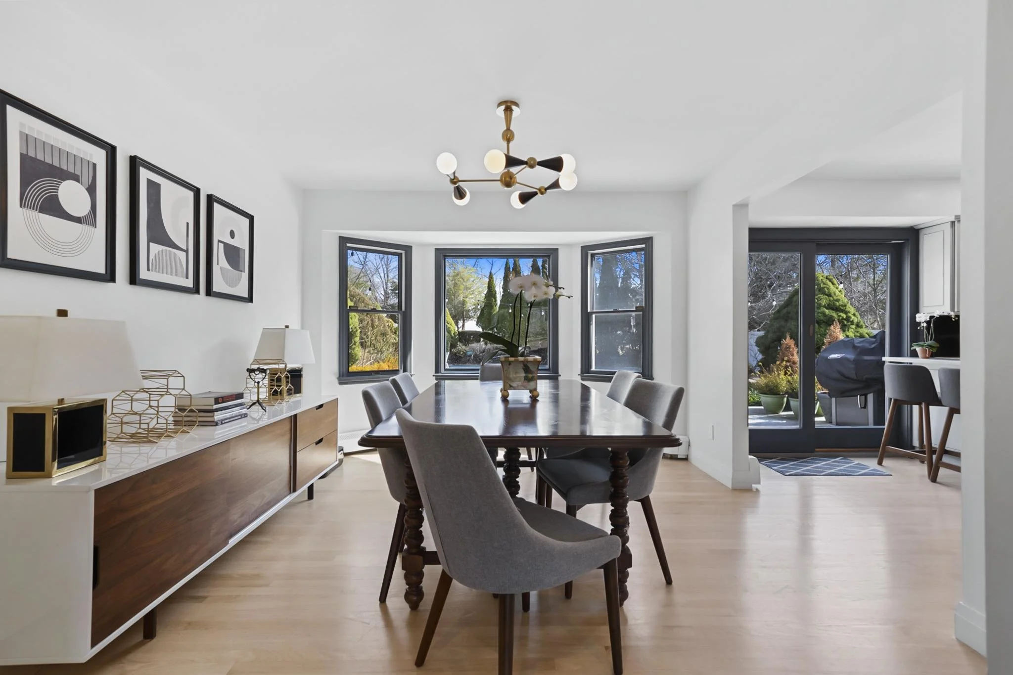 A modern dining room with a large wooden table surrounded by gray upholstered chairs, a white and wooden sideboard with decorative objects, and framed abstract artwork on a white wall. There is a stylish black and brass chandelier overhead. Large bay