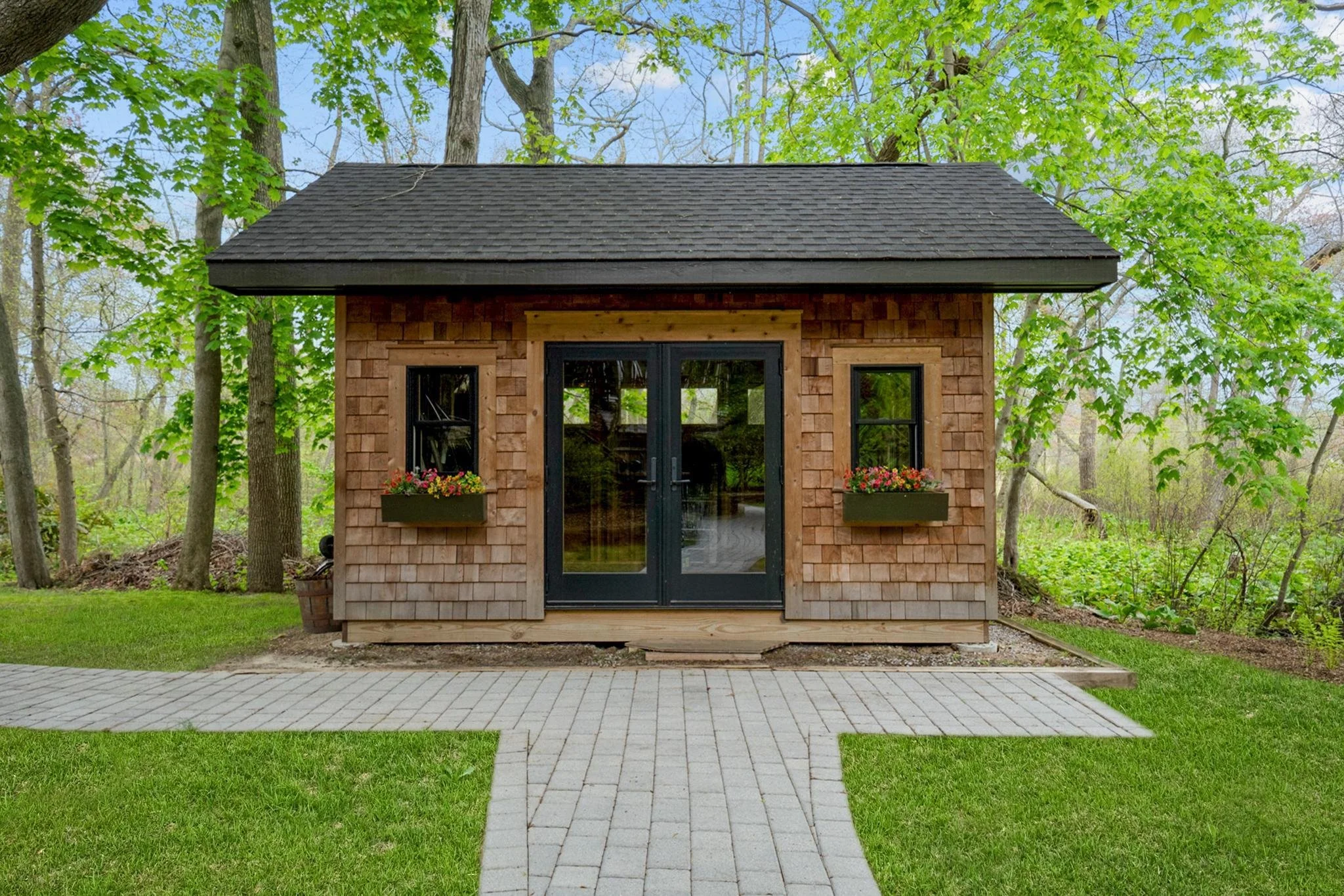 Small wooden house with shingled roof, glass double doors, and two flower boxes on a lush green lawn behind a paved walkway.