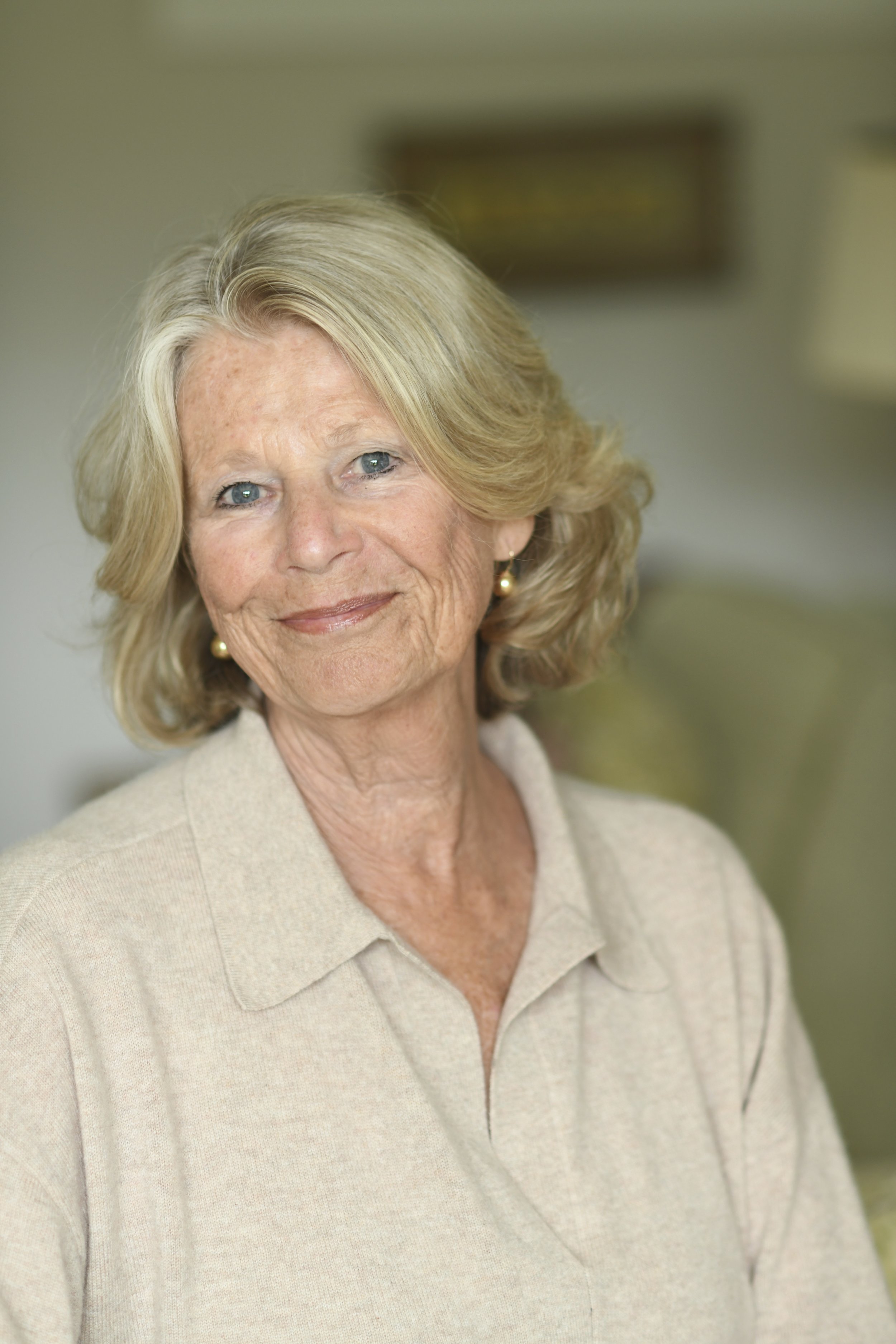 A close-up photograph of an elderly woman with blonde hair, wearing a light beige collared sweater, with blue eyes and earrings, smiling softly. The background is blurred with green and yellow tones.