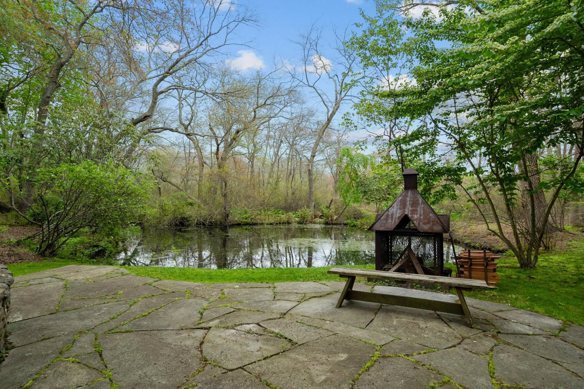 Scenic view of a pond surrounded by trees with fresh green leaves, a small wooden pavilion with a rusted metal roof, a bench, and a paved stone area in the foreground.