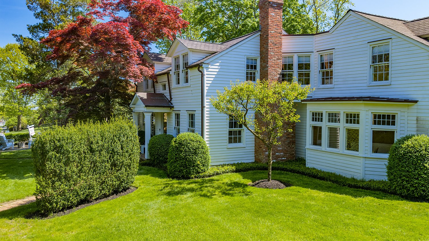 A white two-story house with multiple windows, surrounded by green bushes, trees, and a well-manicured lawn, with a blue sky in the background.