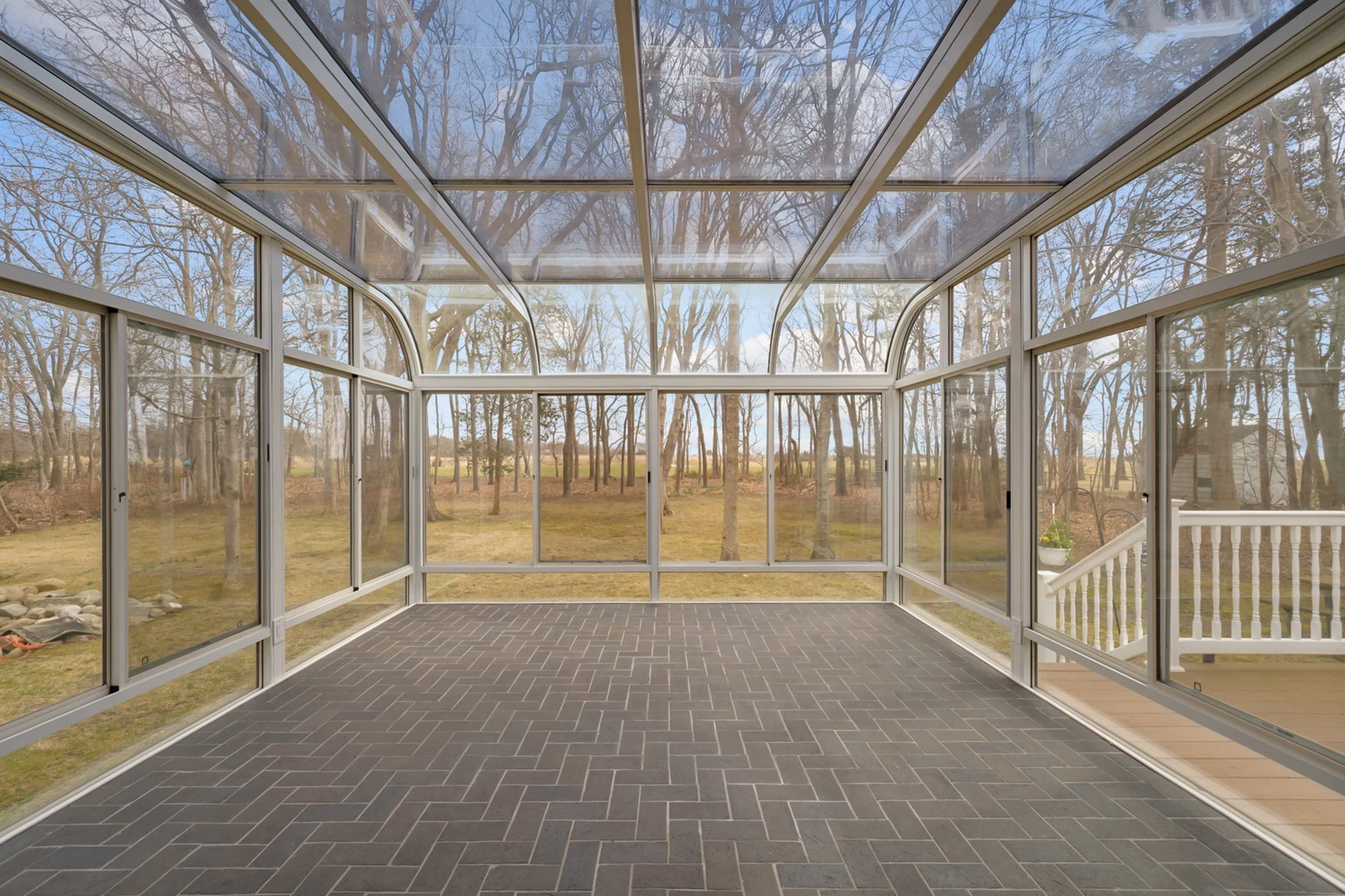 A bright screened-in porch with dark tiled flooring, overlooking a leafless wooded landscape on a clear day.