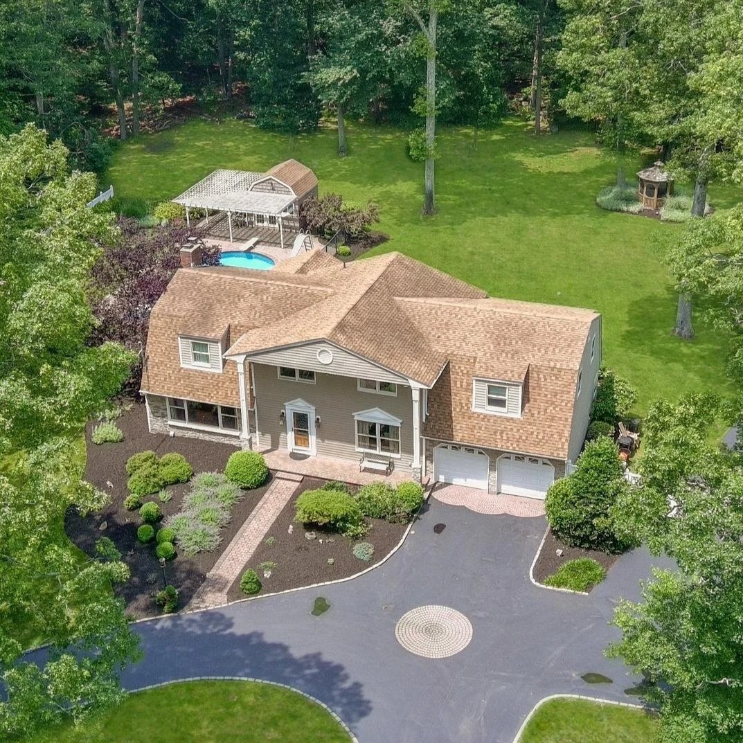 A two-story house with a brown shingled roof, surrounded by a landscaped yard with bushes and trees. There is a driveway with a circular design and a garage. The backyard has a pool, a covered patio, a small shed, and a grassy area with trees.