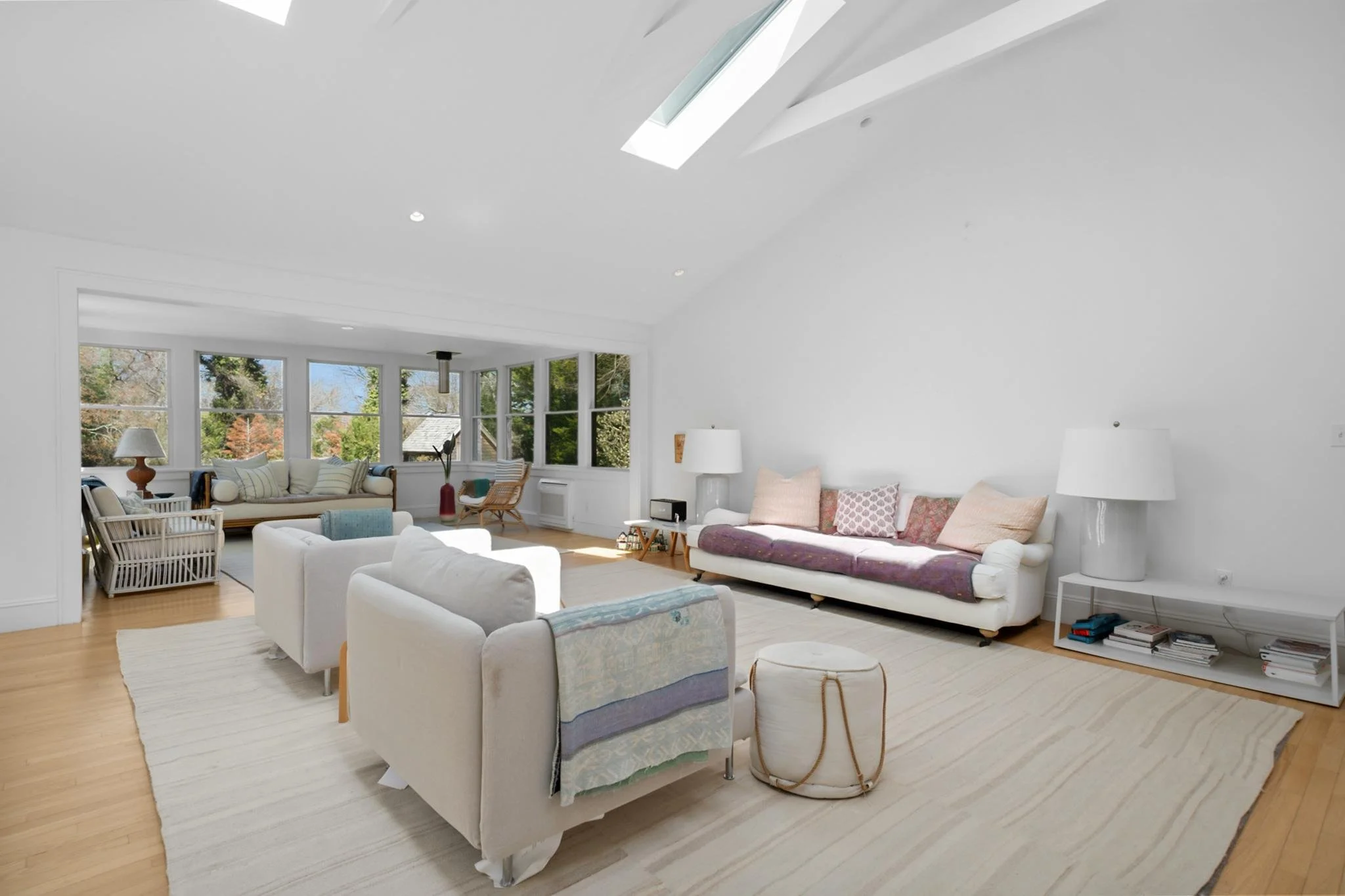 Bright living room with large windows, white walls, and wooden floors, furnished with white sofas, armchairs, side tables, lamps, and decorative pillows.