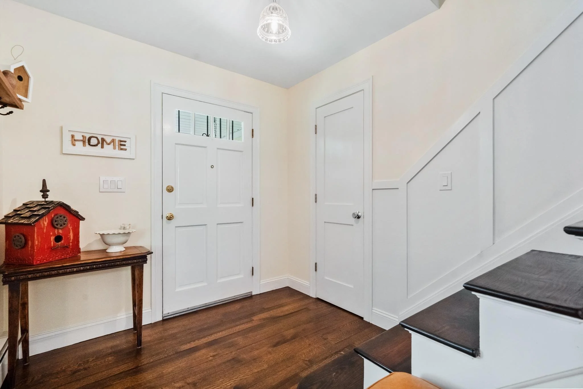 Entryway of a house with a white door with a window, a small wooden table with a red birdhouse and a white decorative bowl, a chalkboard sign with the word 'Home,' and stairs with black treads and white risers.