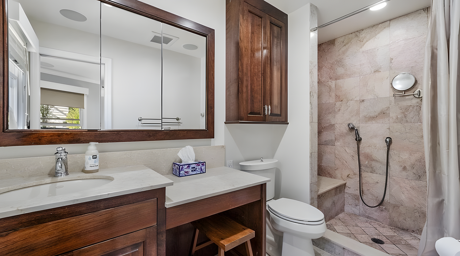 Bathroom with wooden cabinets, a large mirror, a white countertop with a sink, a tissue box, a toilet, and a shower area with beige marble tiles, a built-in bench, and a handheld shower head.