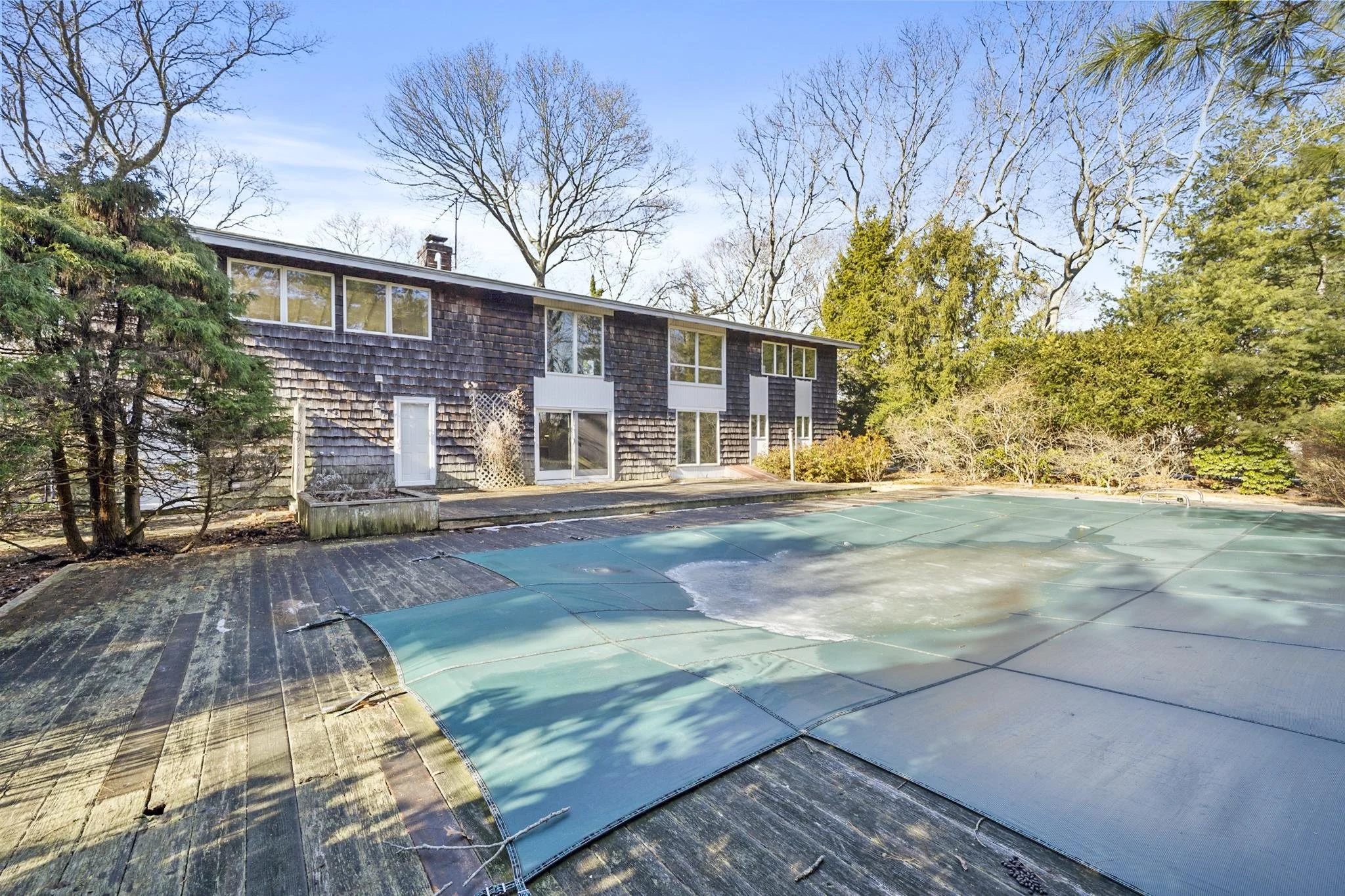 Backyard scene with a house featuring shingles, leafless trees in the background, and an empty swimming pool with a green cover in the foreground.