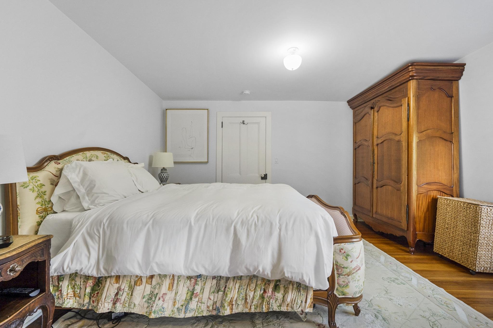 A bedroom with a large bed covered in white linens and floral bedding, wooden nightstands, a lamp, framed artwork on the wall, a wooden wardrobe, and a chair with a woven back, with hardwood floors and marble-patterned area rug.