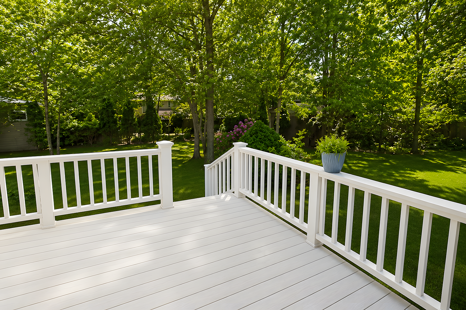 View of a backyard with a white deck and railing, green grass, trees, and purple flowering bush in the background, with a potted plant on the deck railing.