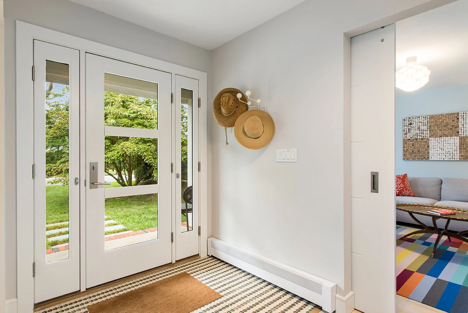Entryway with white front door, two small window panels on each side, and door to living room. Hats hanging on wall hooks. View of garden through door windows. Living room with colorful rug, couch, and wall art visible through an open doorway.