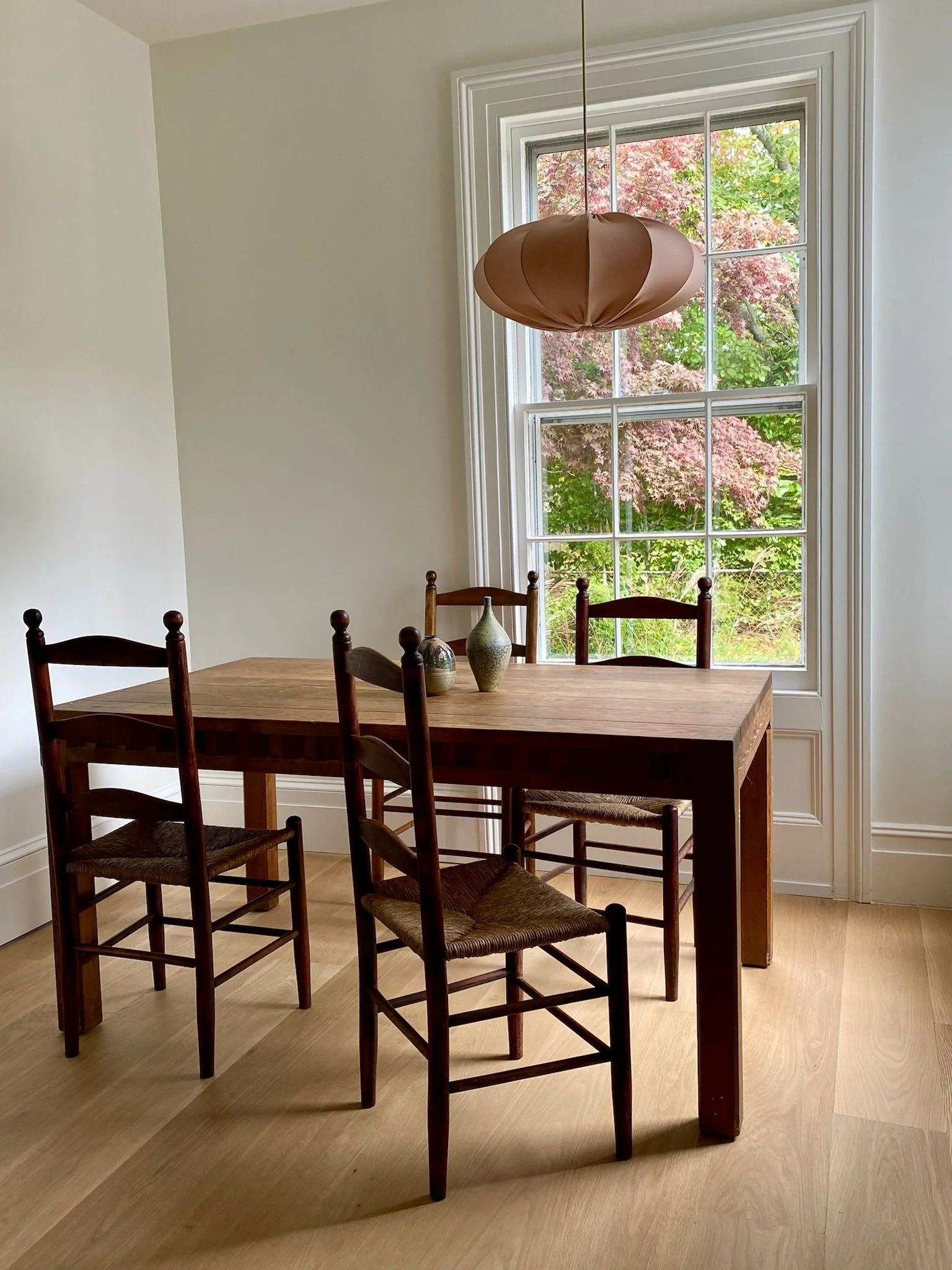 A dining room with a wooden table and six wooden chairs with woven seats, three on each side. There are three decorative vases in different shapes and colors on the table. A large window with white trim shows pink and green trees outside. A pink pend
