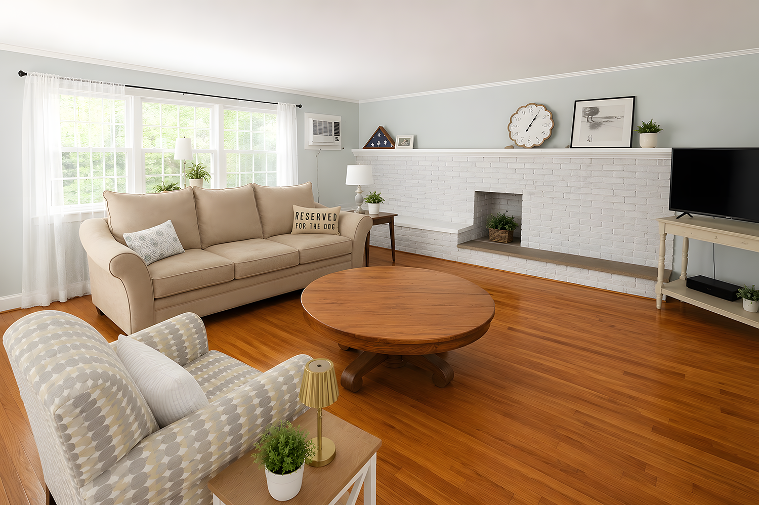 Living room with a beige sofa, a white floral armchair, a round wooden coffee table, and a white brick fireplace with decorative items, a wall clock, and a window with white curtains.