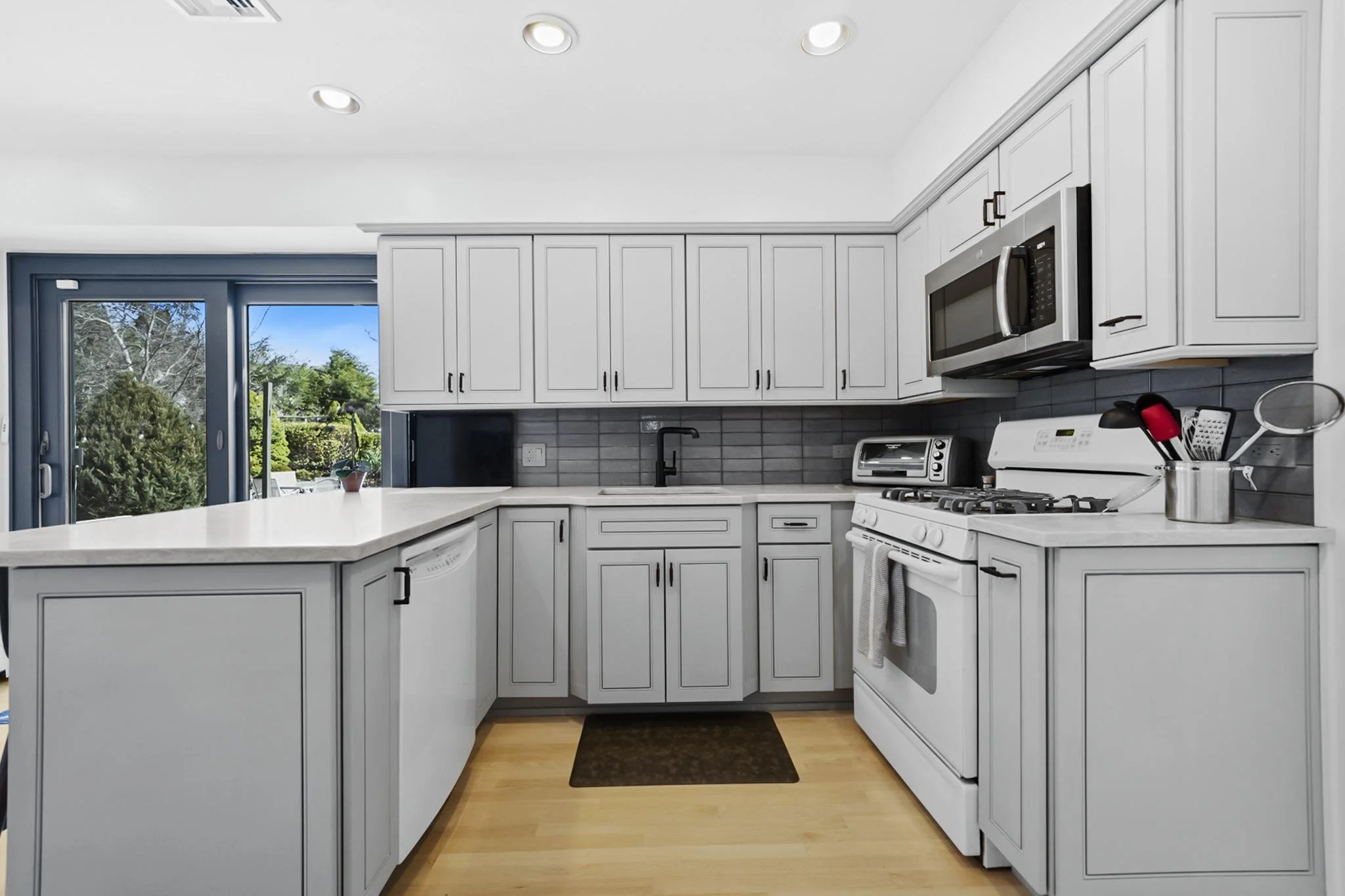 Kitchen with white cabinets, black hardware, grey tile backsplash, black faucet, white appliances, black utensil holder, and a sliding glass door showing outdoor greenery.