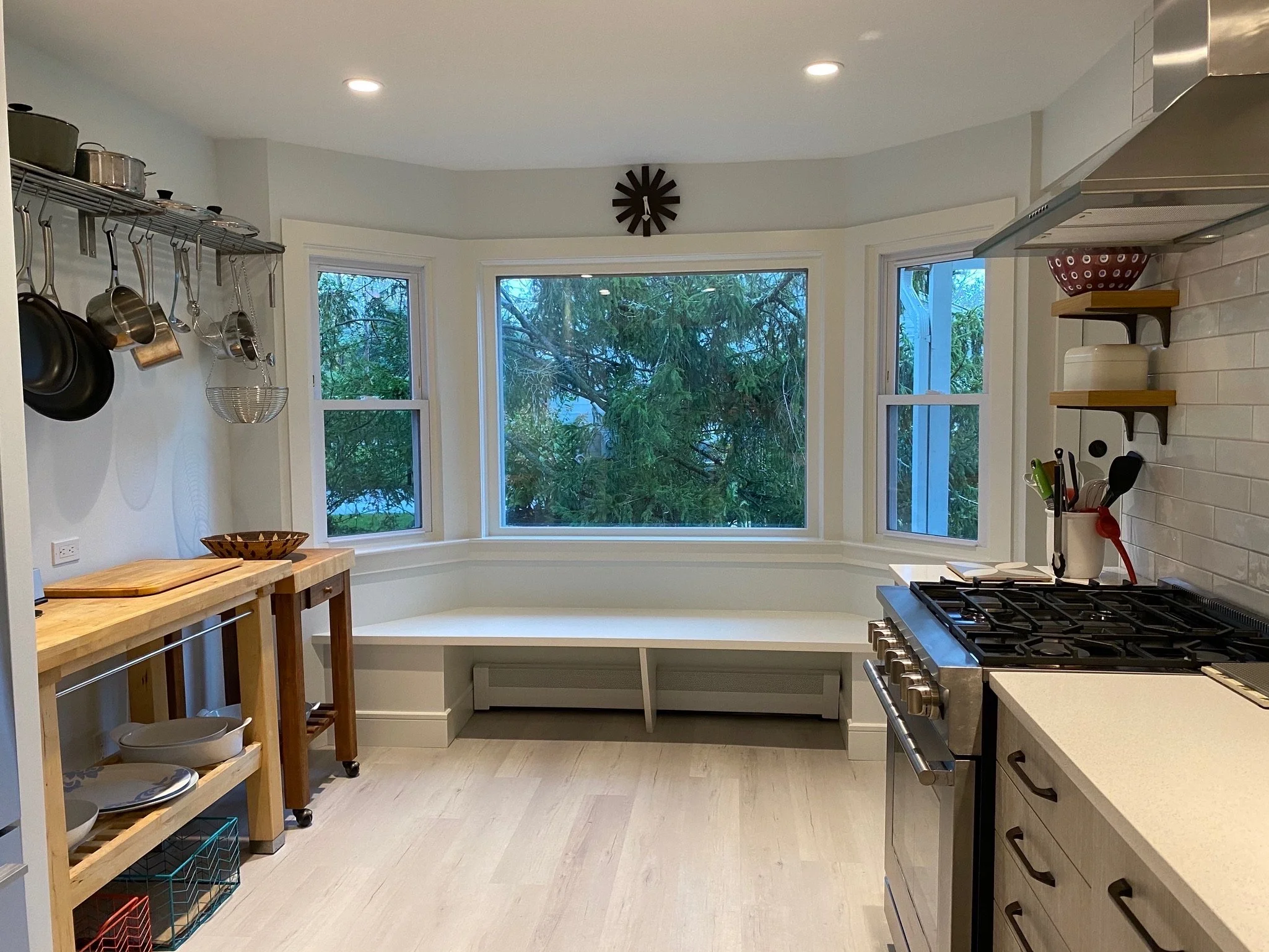 Kitchen with large window overlooking trees, stainless steel stove, wooden shelves with bowls and utensils, hanging pots and pans, and a clock on the wall.