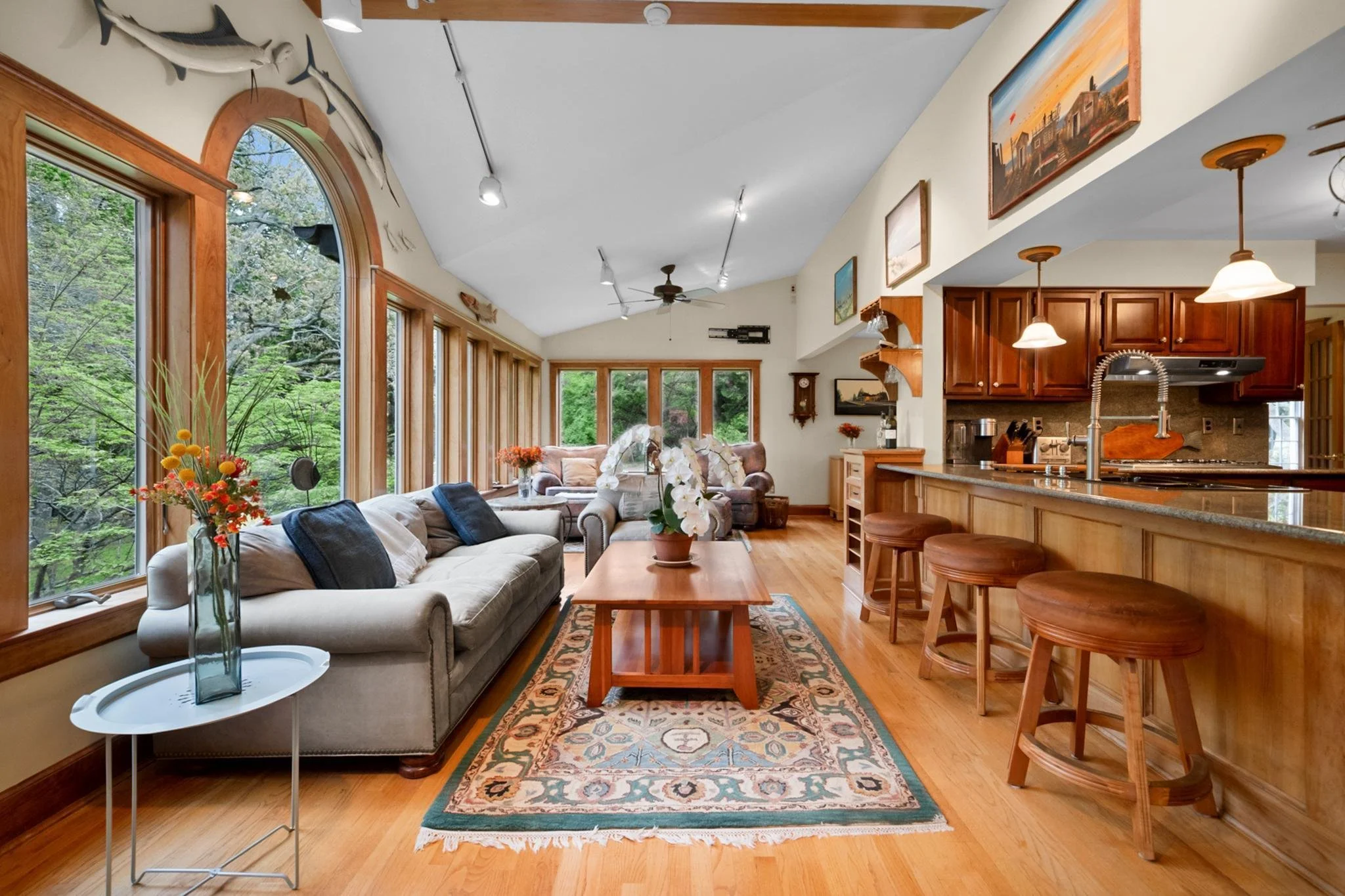 Living room with large windows overlooking trees, furnished with a beige sofa, armchairs, a wooden coffee table, and decorated with plants and flowers, with wood flooring and artwork on the walls.