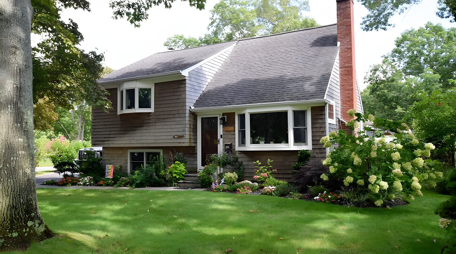 A two-story house with brown siding and white-framed windows, surrounded by a lush green garden with various flowers and a well-maintained lawn.