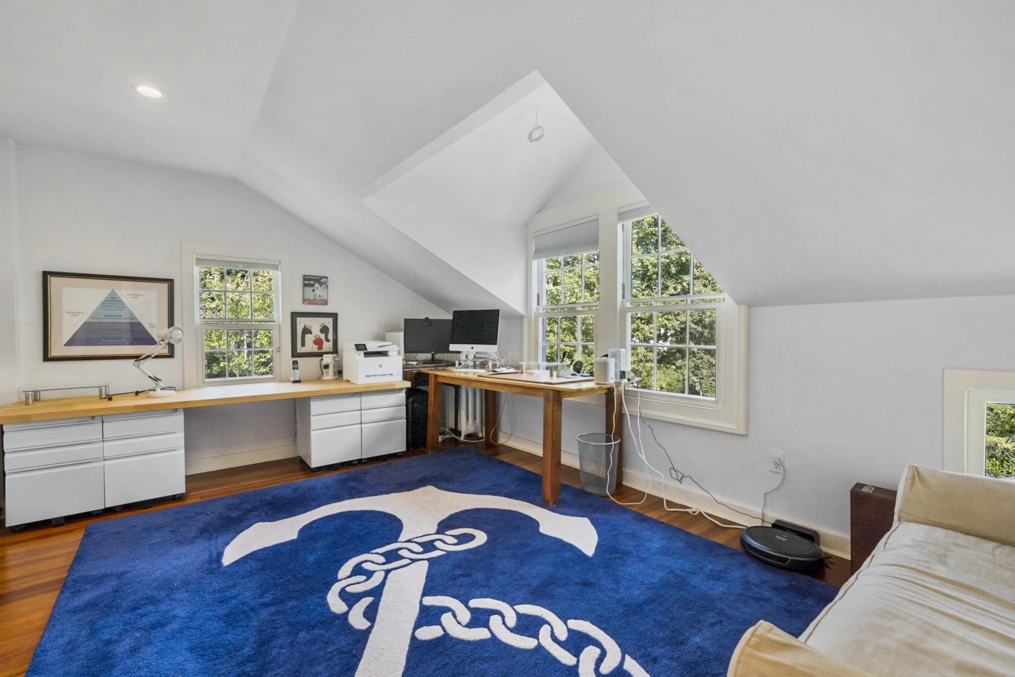 A bright attic room with white walls and large windows, a desk with a computer, printer, and office supplies, a blue rug with a white logo, and a beige sofa.