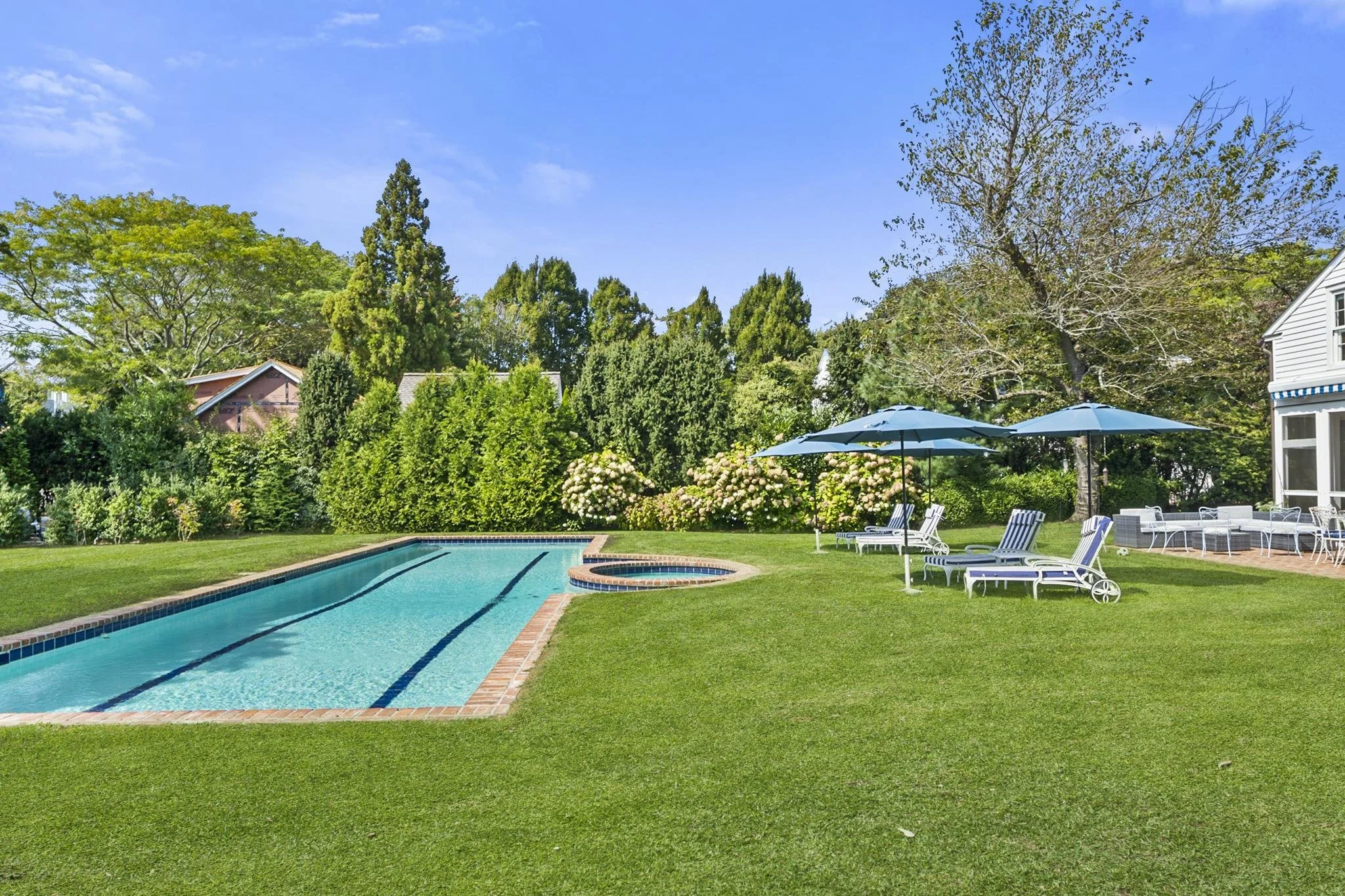 Backyard with swimming pool, lounge chairs, umbrellas, and lush green trees under a clear blue sky.